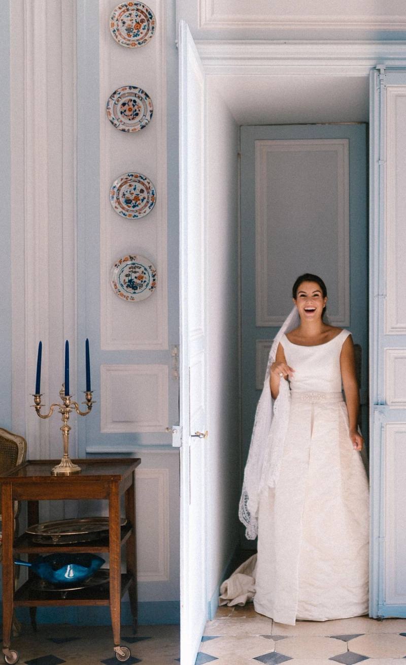 Smiling bride with a long veil stepping through a doorway, a candelabra and wall plates beside her