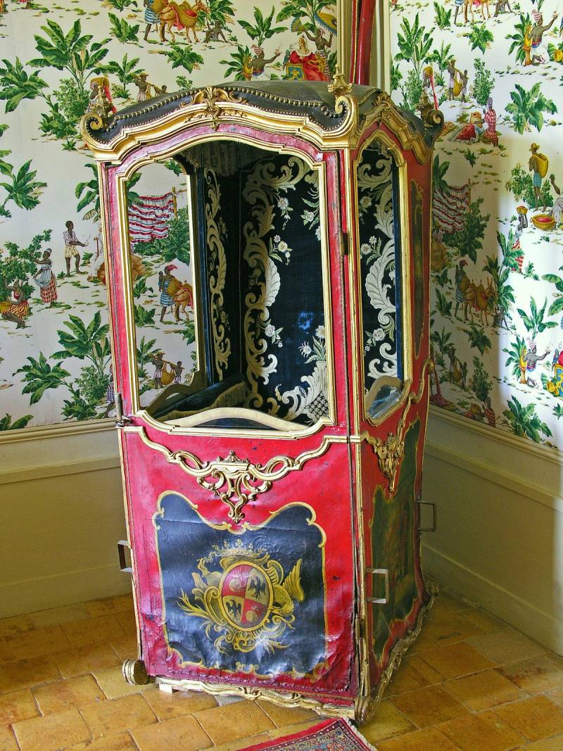 Chaise à porteurs ancienne rouge et dorée, ornée d'armoiries, devant un papier peint à scènes exotiques