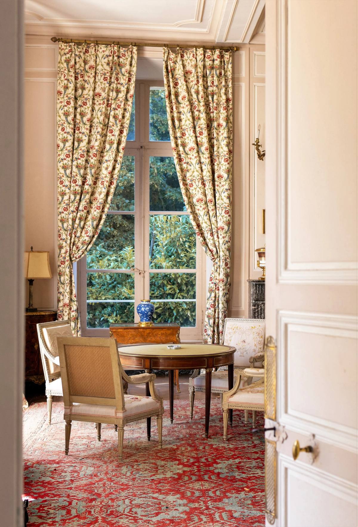 The Grand Salon glimpsed through an open door: tall window, floral curtains, round table and red rug