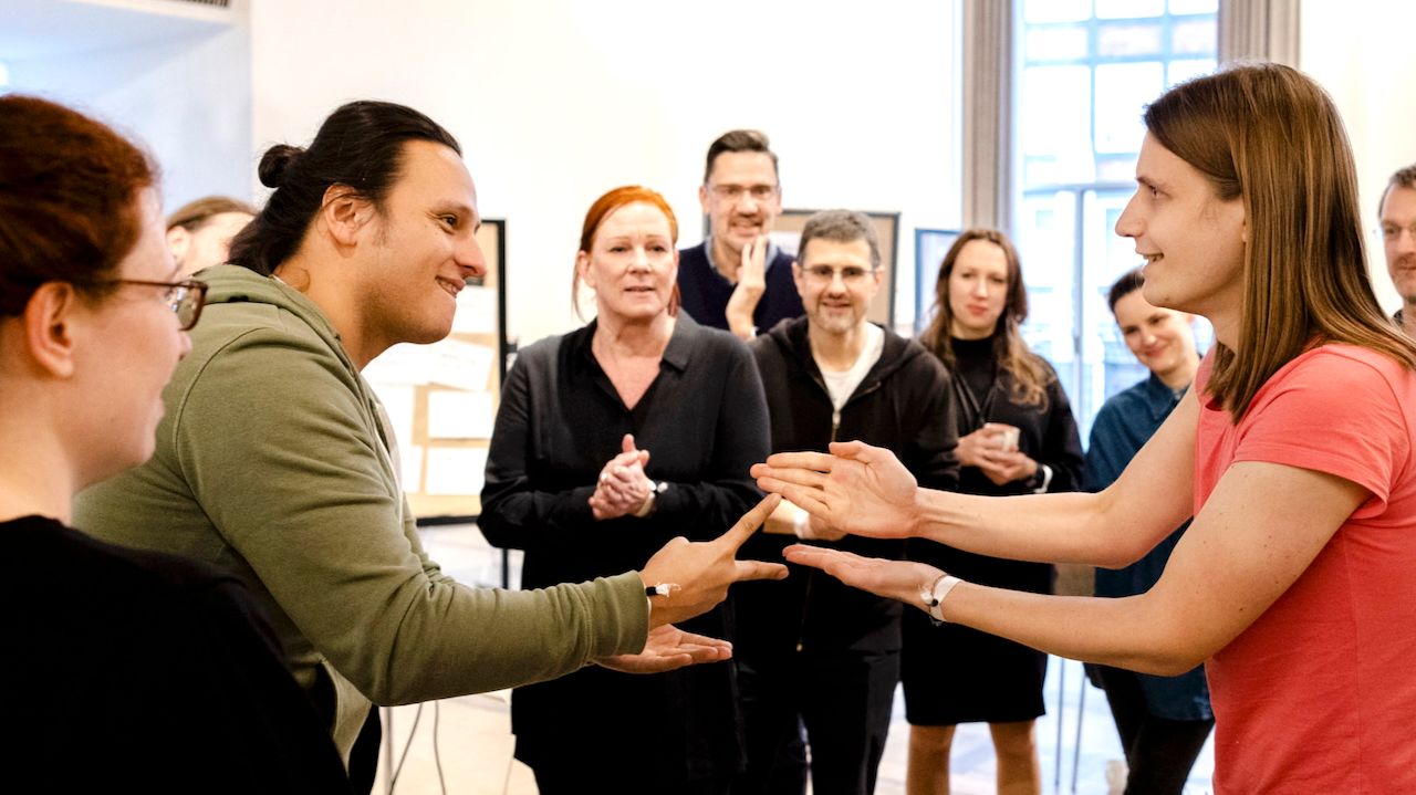 Zwei Teammitglieder spielen Schere-Stein-Papier, umgeben von applaudierenden Kolleg:innen – Symbol für gemeinsames Entscheiden und Teamstärkung