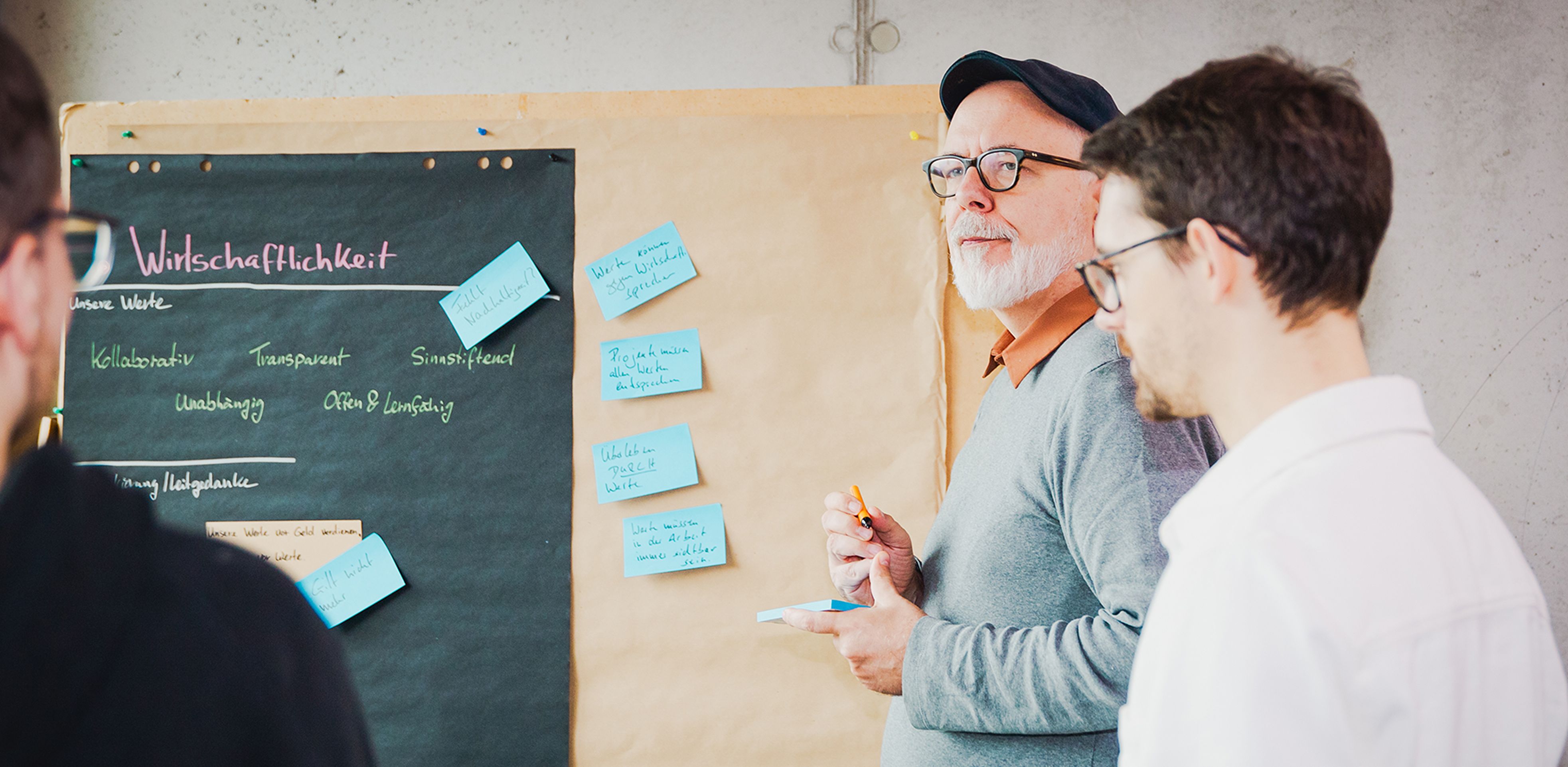 Zwei Softwareentwickler diskutieren mit einem Kunden vor einer Moderationswand über die Produktentwicklung.