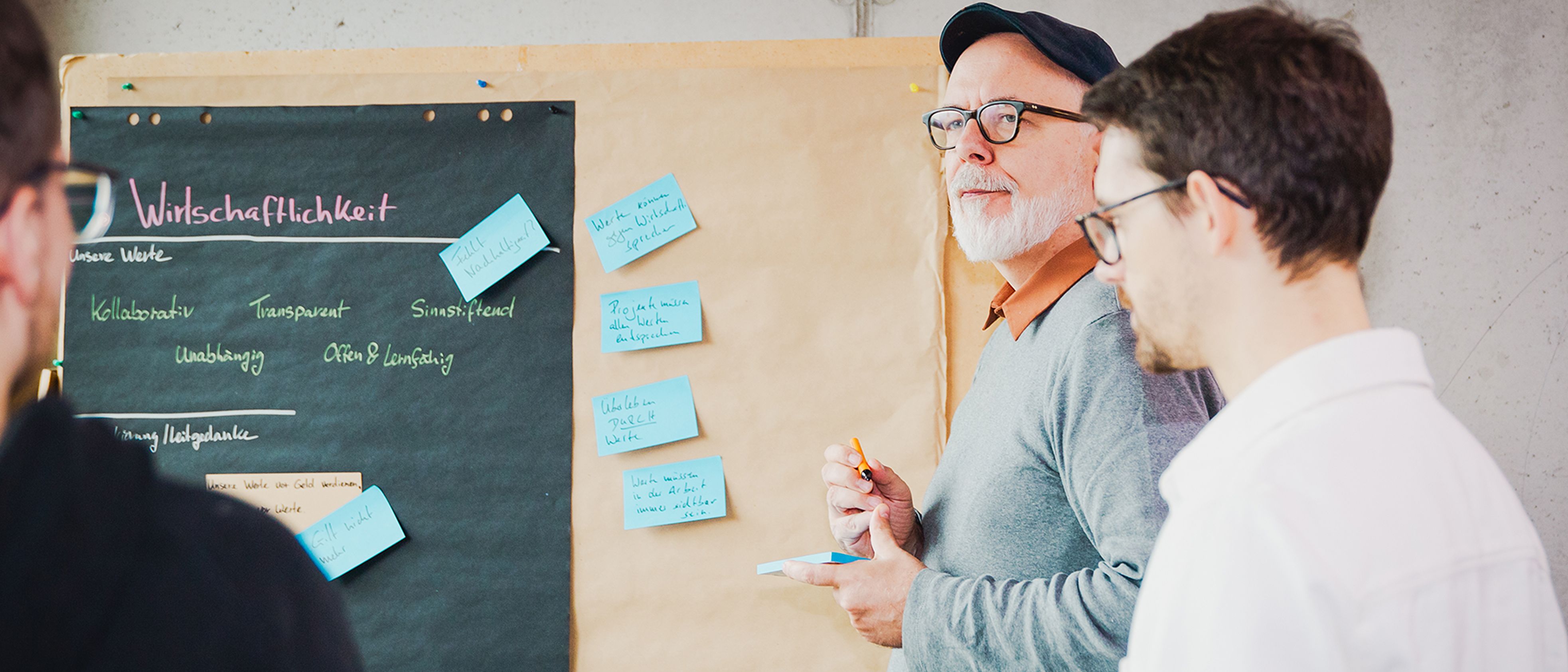 Kooperative Softwareentwicklung mit externen Teams  Zwei Softwareentwickler diskutieren mit einem Kunden vor einer Moderationswand über die Produktentwicklung.