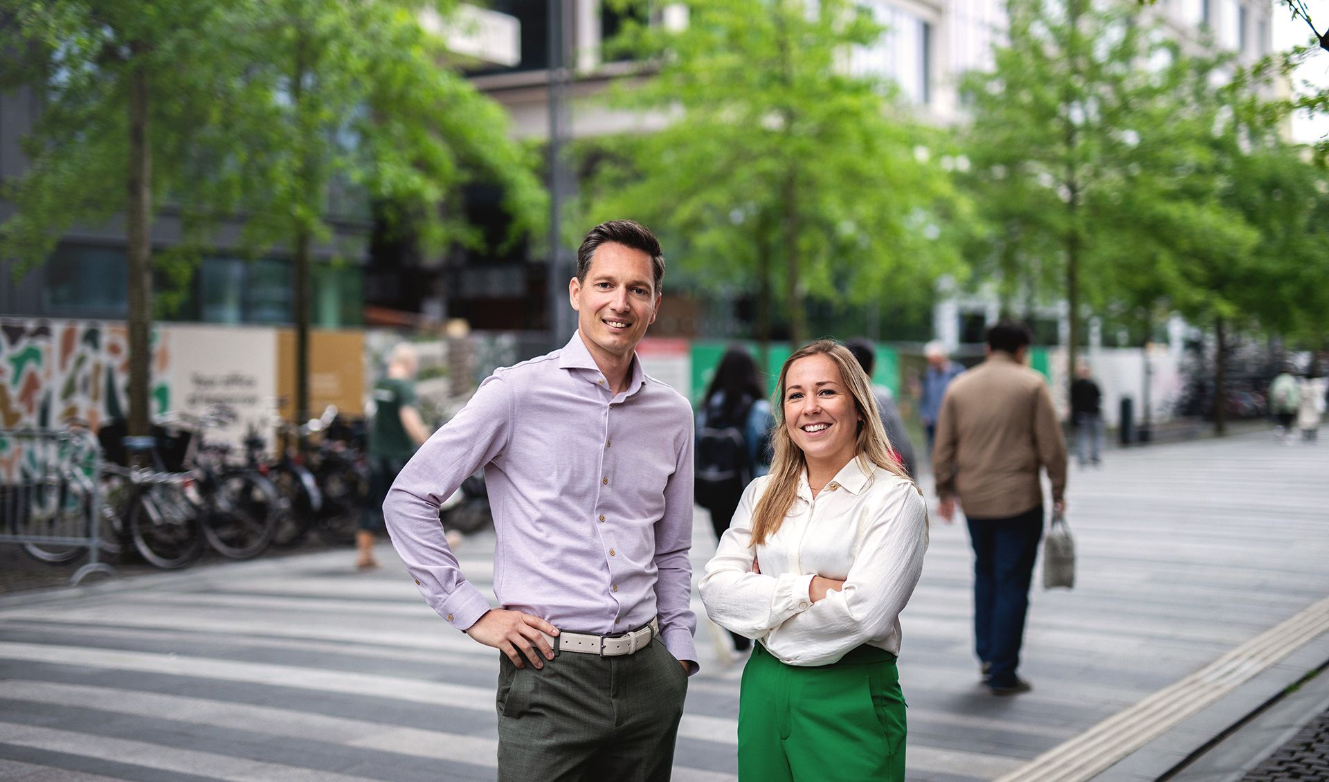 Twee mensen, een man in een lichtpaarse shirt en een vrouw in een groene broek, staan op een voetgangersvriendelijke straat met bomen en geparkeerde fietsen. Andere mensen lopen voorbij op de achtergrond.