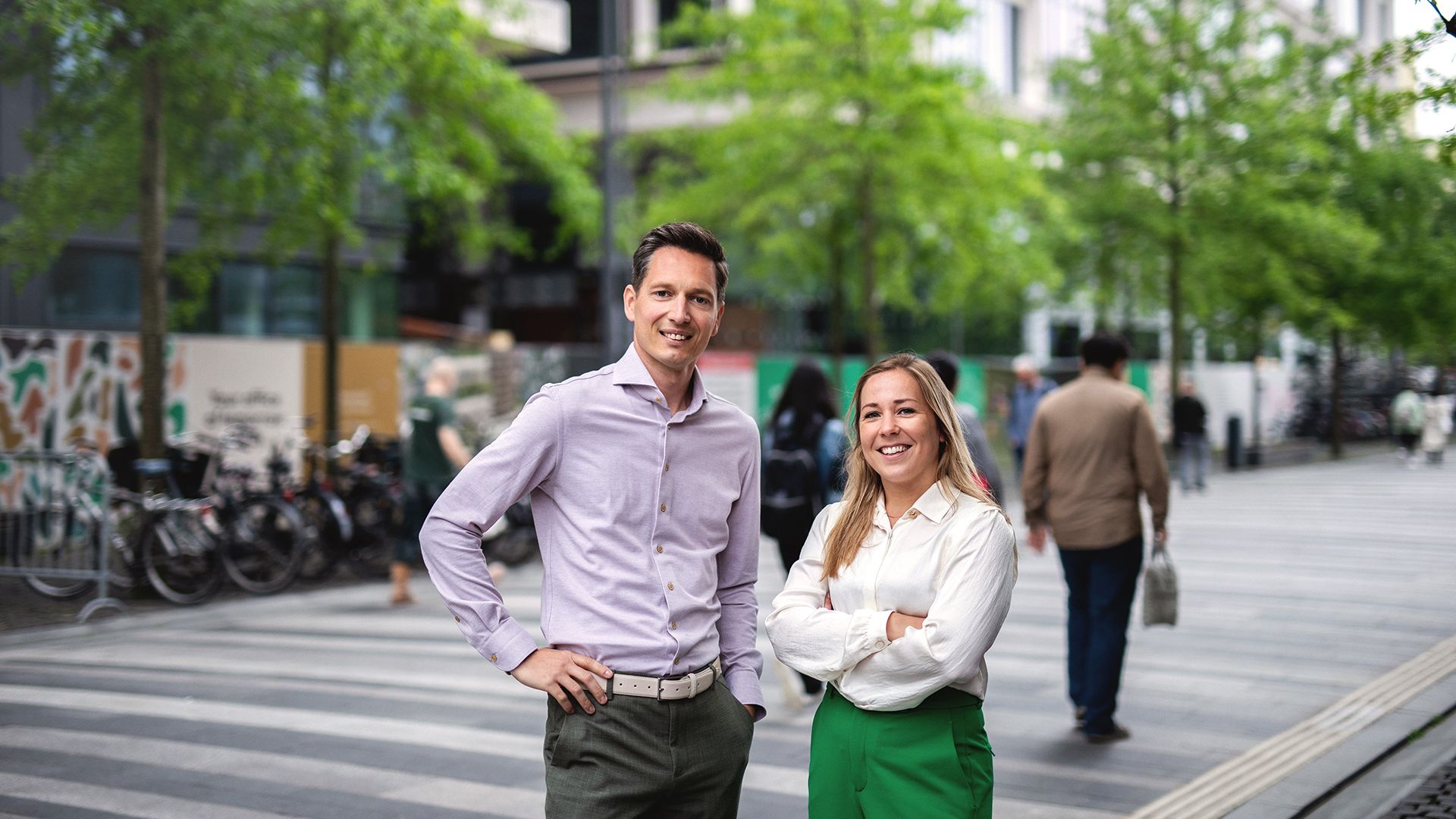 Twee mensen, een man in een lichtpaarse shirt en een vrouw in een groene broek, staan op een voetgangersvriendelijke straat met bomen en geparkeerde fietsen. Andere mensen lopen voorbij op de achtergrond.
