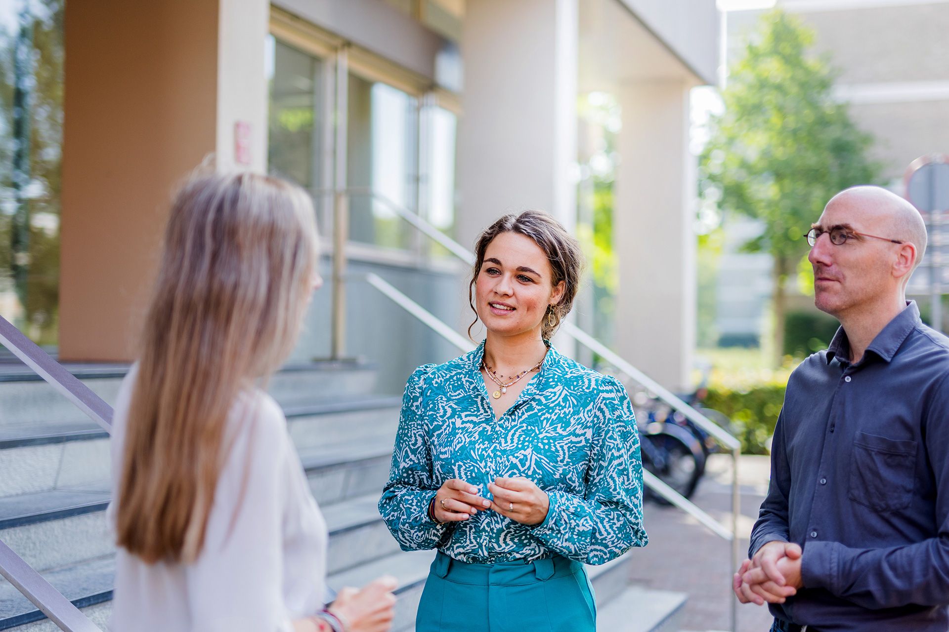 Vrouw in blauwe blouse praat buiten met twee collega's voor een gebouw.