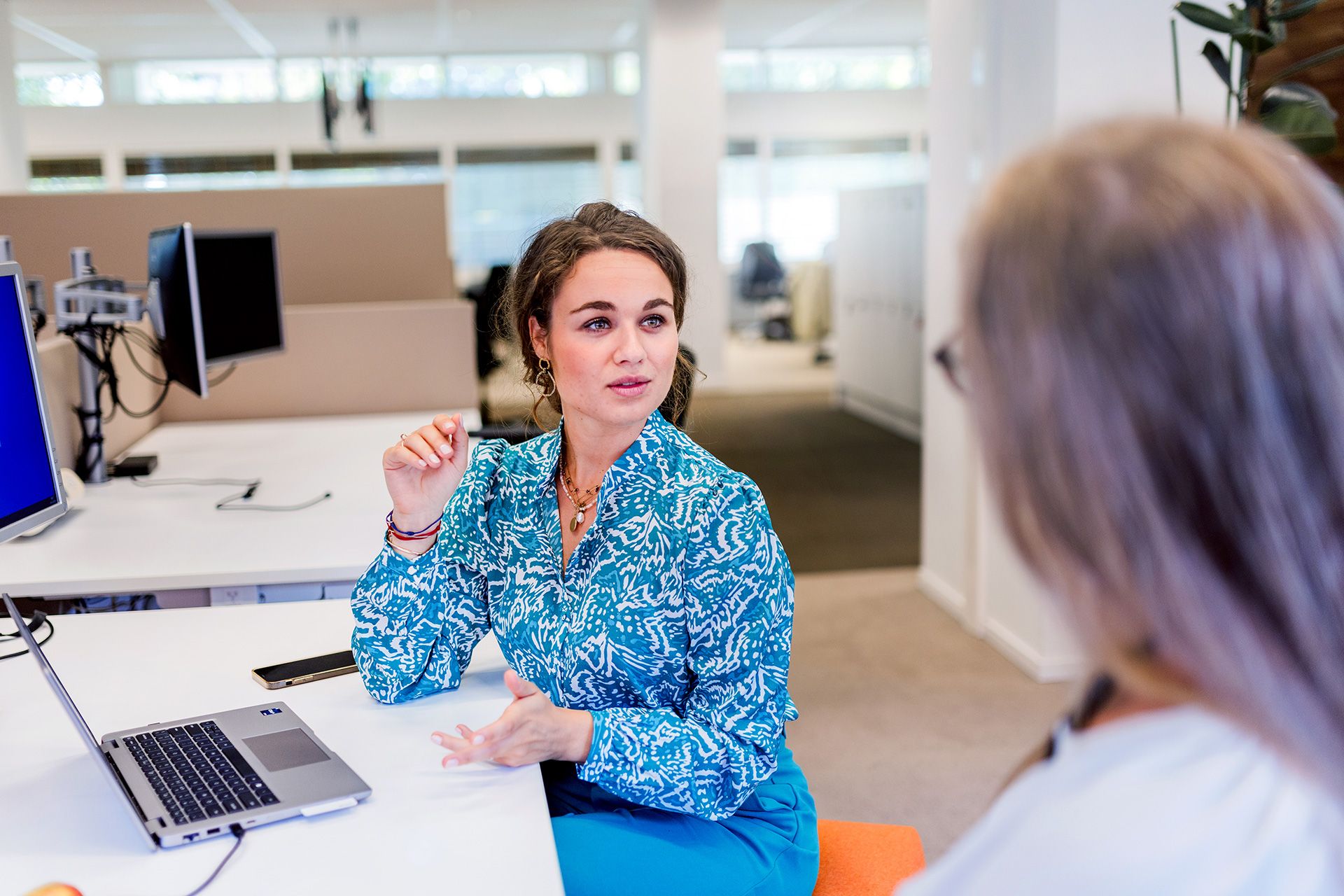 Vrouw in blauwe blouse praat met een collega aan een bureau in een kantoor.