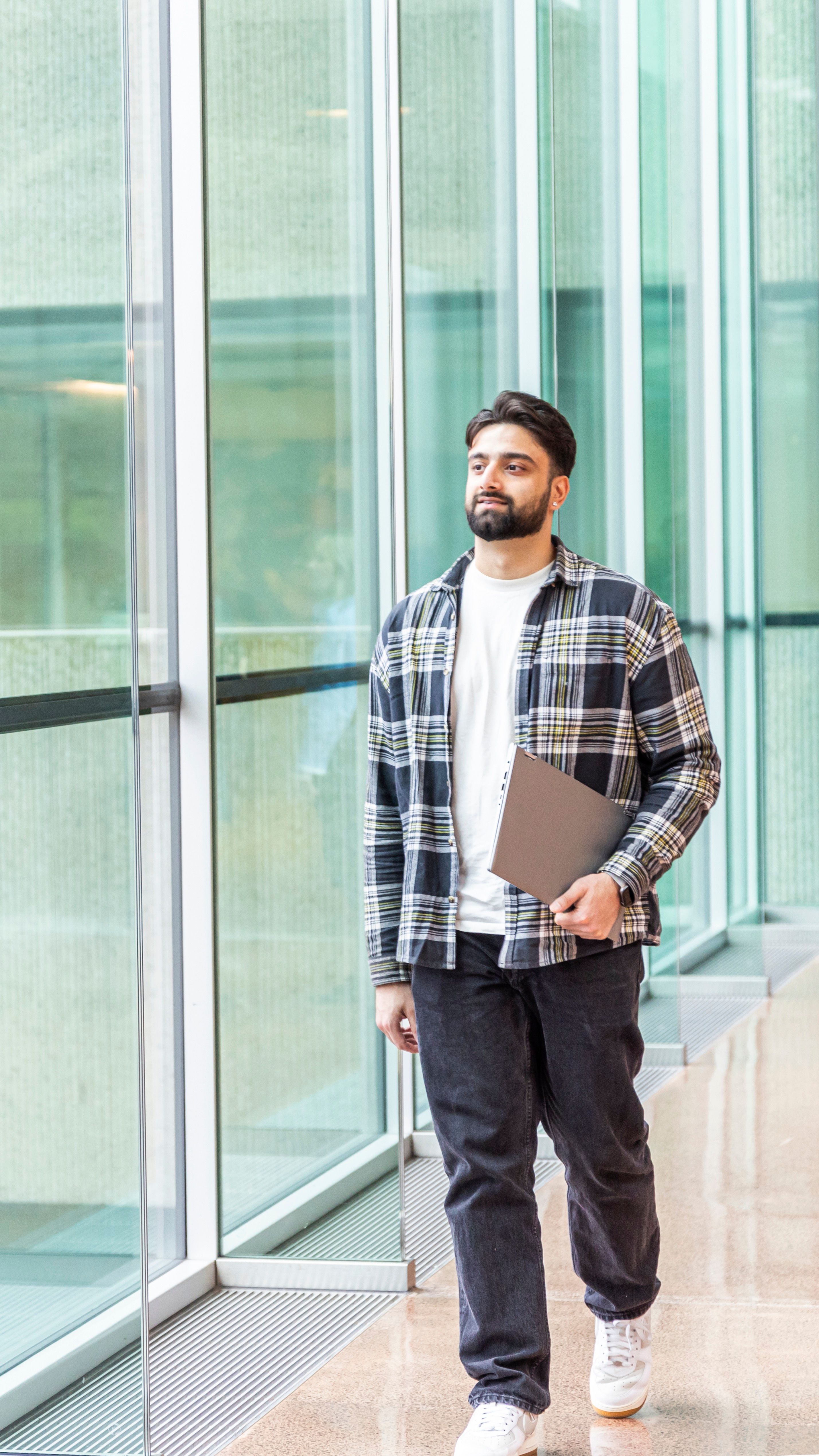 A man in a plaid shirt and jeans standing thoughtfully in a brightly lit space with large glass windows, holding a laptop by his side.