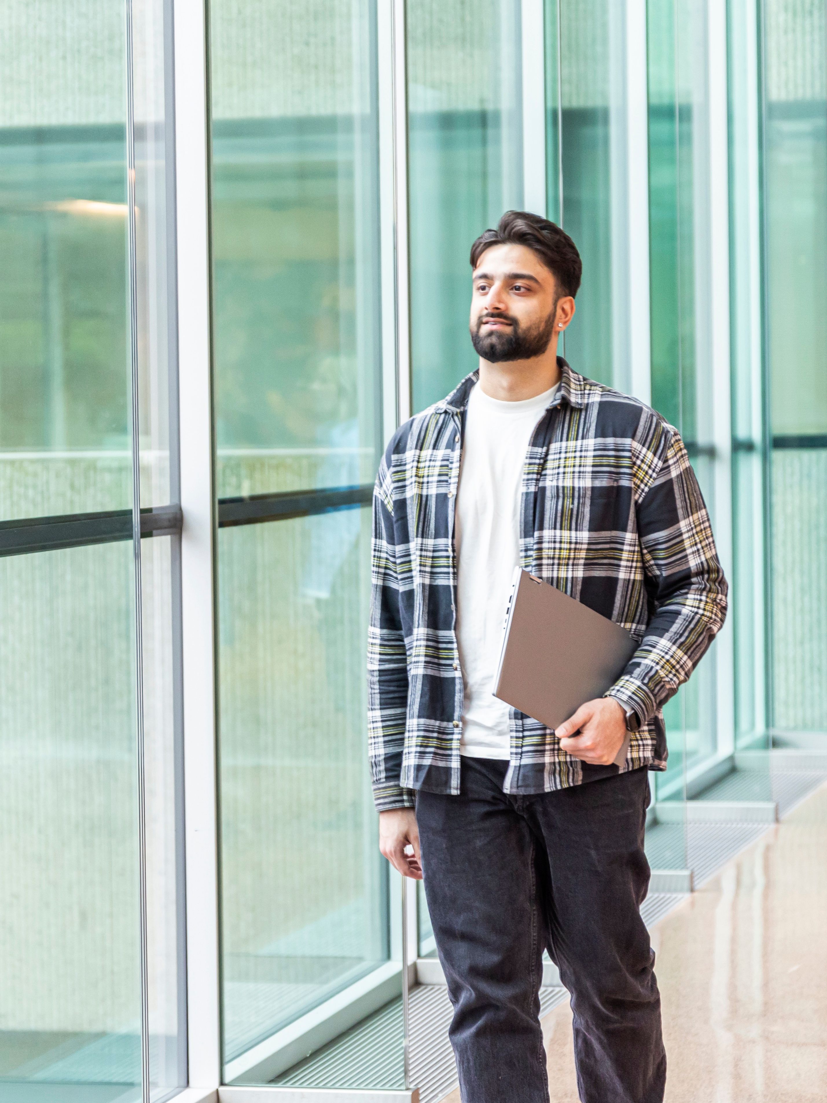 A man in a plaid shirt and jeans standing thoughtfully in a brightly lit space with large glass windows, holding a laptop by his side.