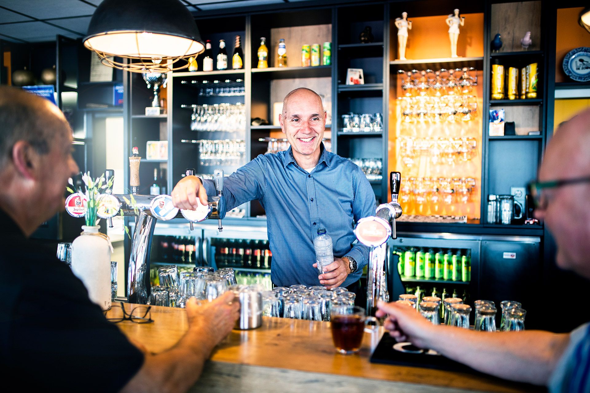 Sjaak Staal achter de bar en leunt tegen biertap. Twee mannen zitten aan de bar