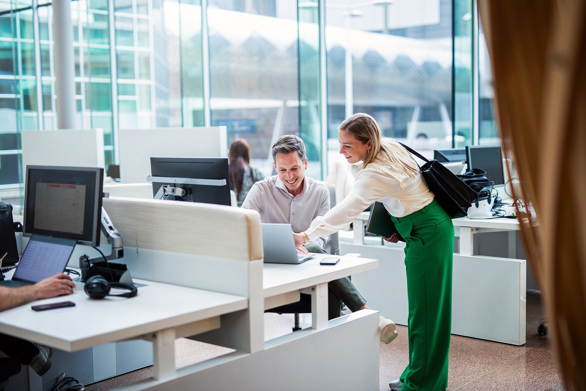  Een man en een vrouw staan in een moderne kantoorruimte. De vrouw wijst naar het scherm van de laptop van de man. De kantoorruimte heeft grote ramen en meerdere werkstations.