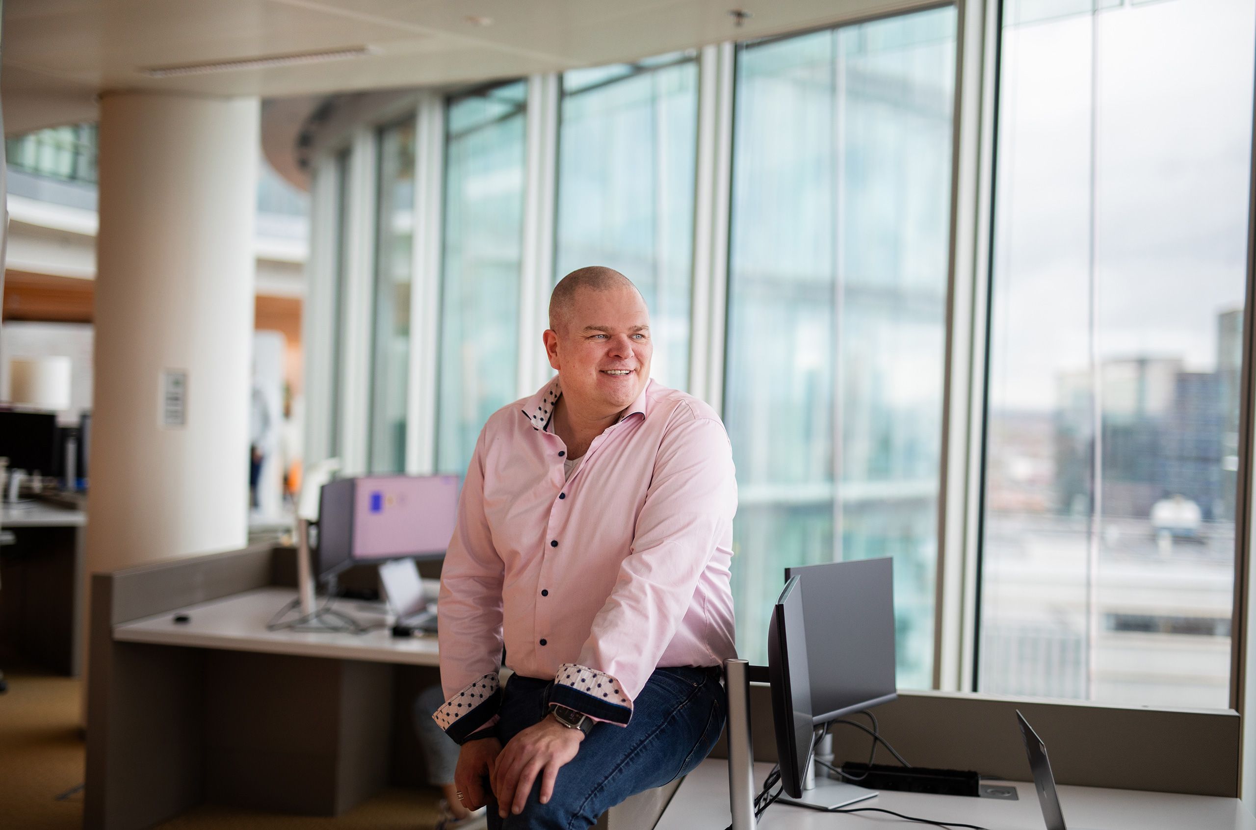 Person in pink shirt smiling at modern office desk with large windows overlooking cityscape, computer monitors nearby.