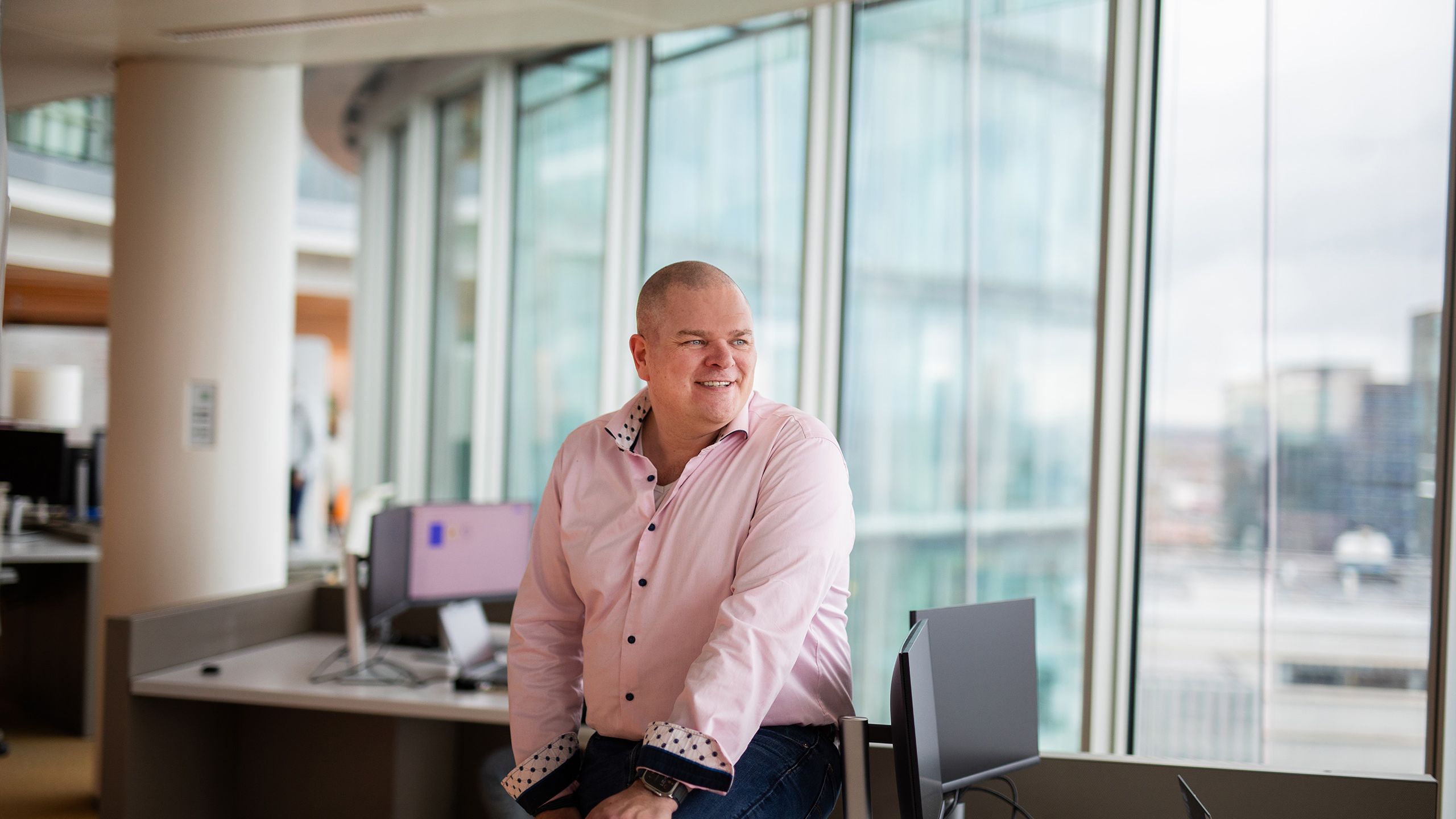 Person in pink shirt smiling at modern office desk with large windows overlooking cityscape, computer monitors nearby.