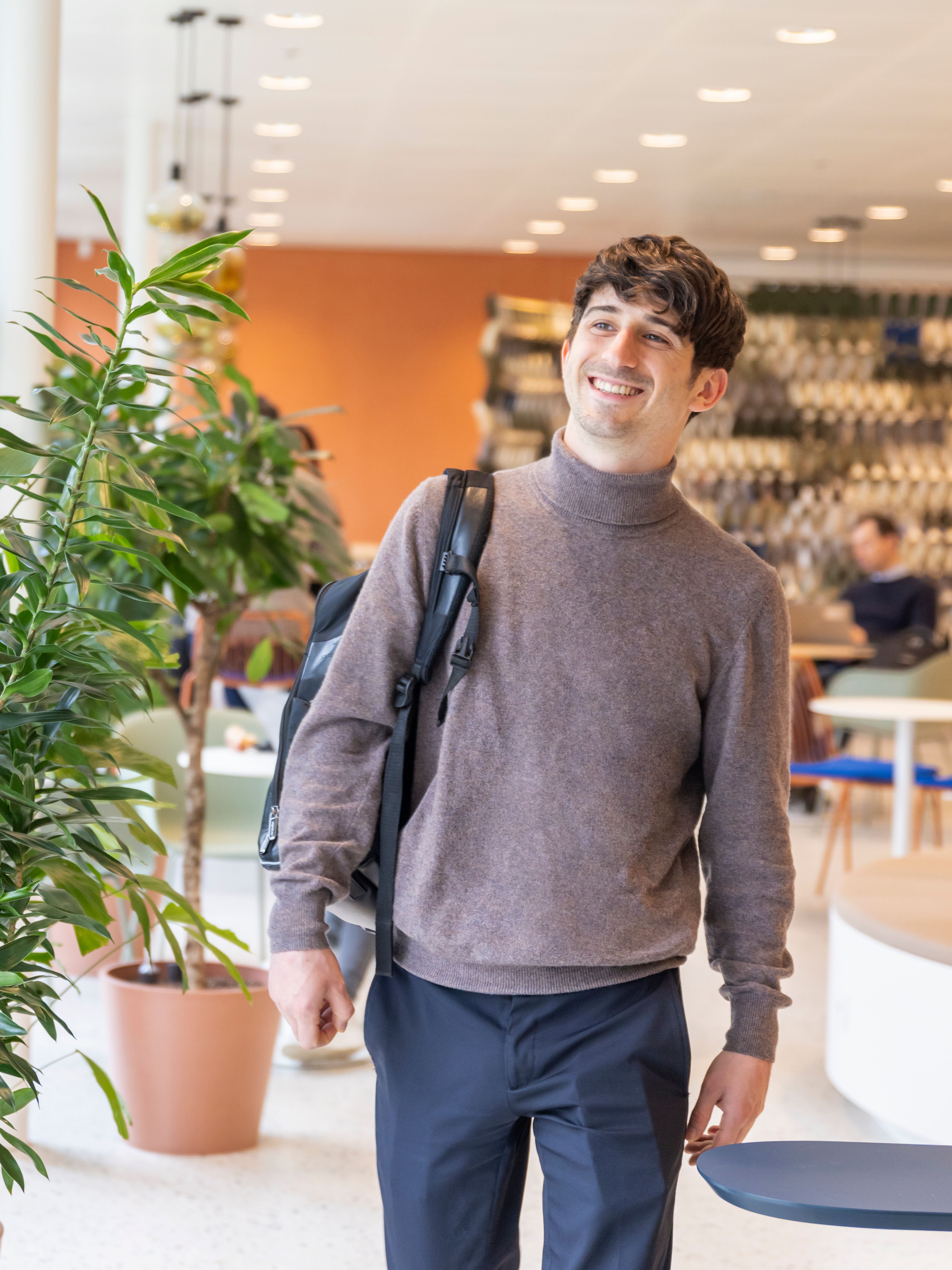A smiling young man in a grey turtleneck and navy pants walking in a bright indoor setting with modern furniture and a potted plant.