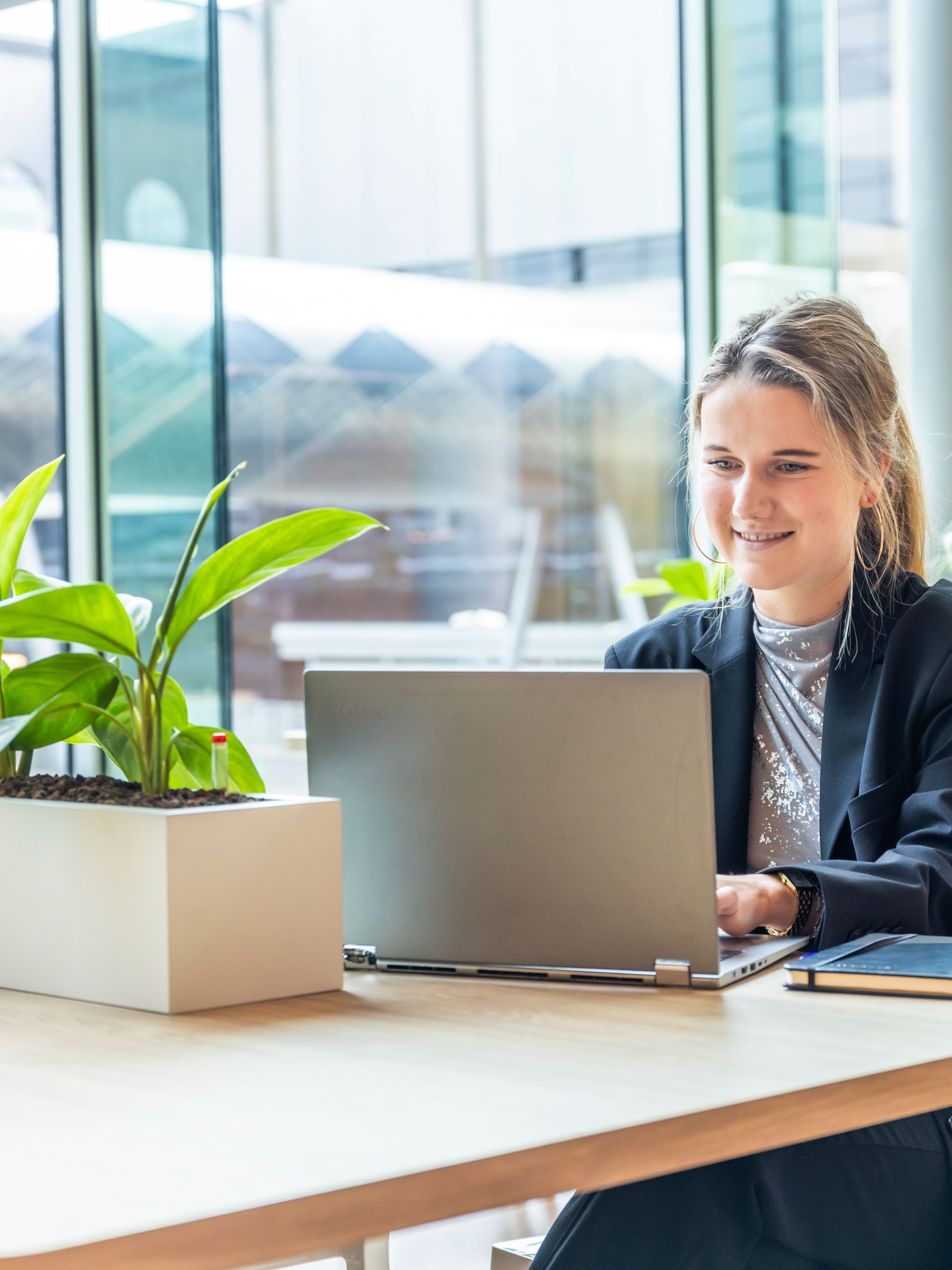 A smiling young woman in professional attire working on a laptop at a desk in a well-lit office, with a notebook and smartphone beside her.
