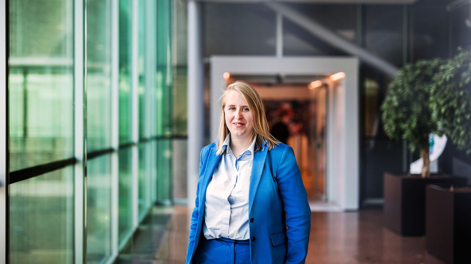 Woman in blue suit (Business Controller) standing at glass wall.