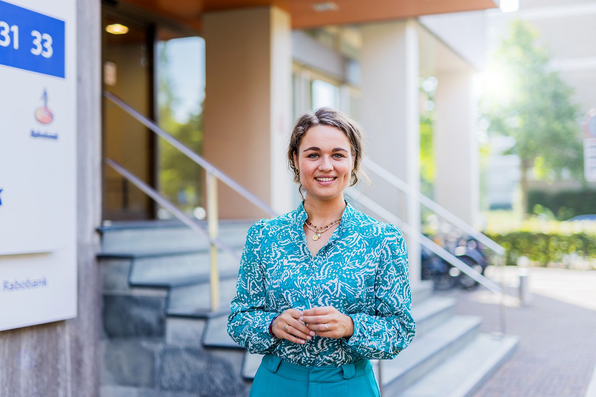 Een vrouw in een blauw patroonblouse staat buiten een gebouw en glimlacht recht in de camera. Ze staat bij een trap met een gebouwingang en wat groen op de achtergrond.