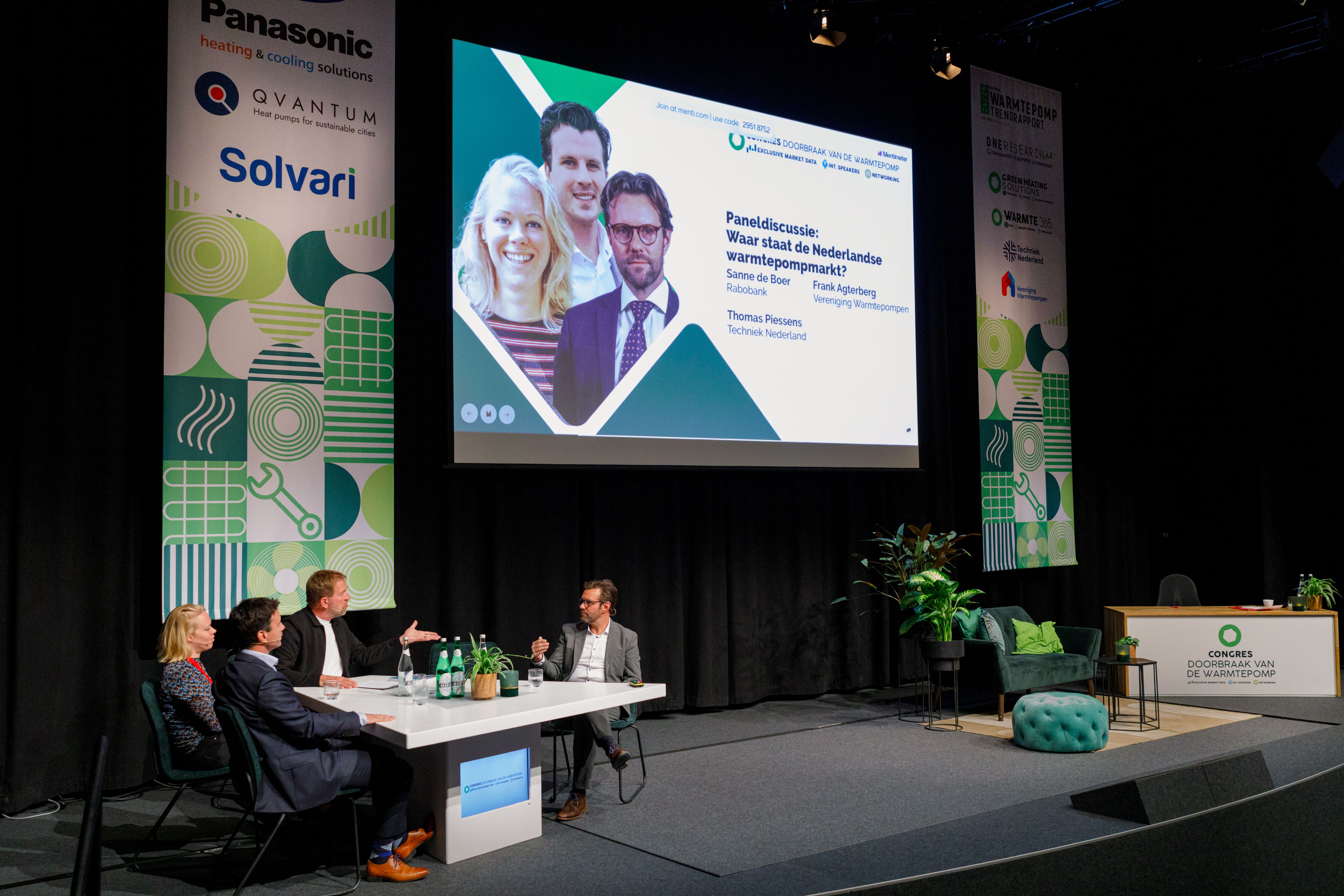 Een vrouw en 3 mannen zitten aan een tafel en zijn in gesprek met elkaar op een podium