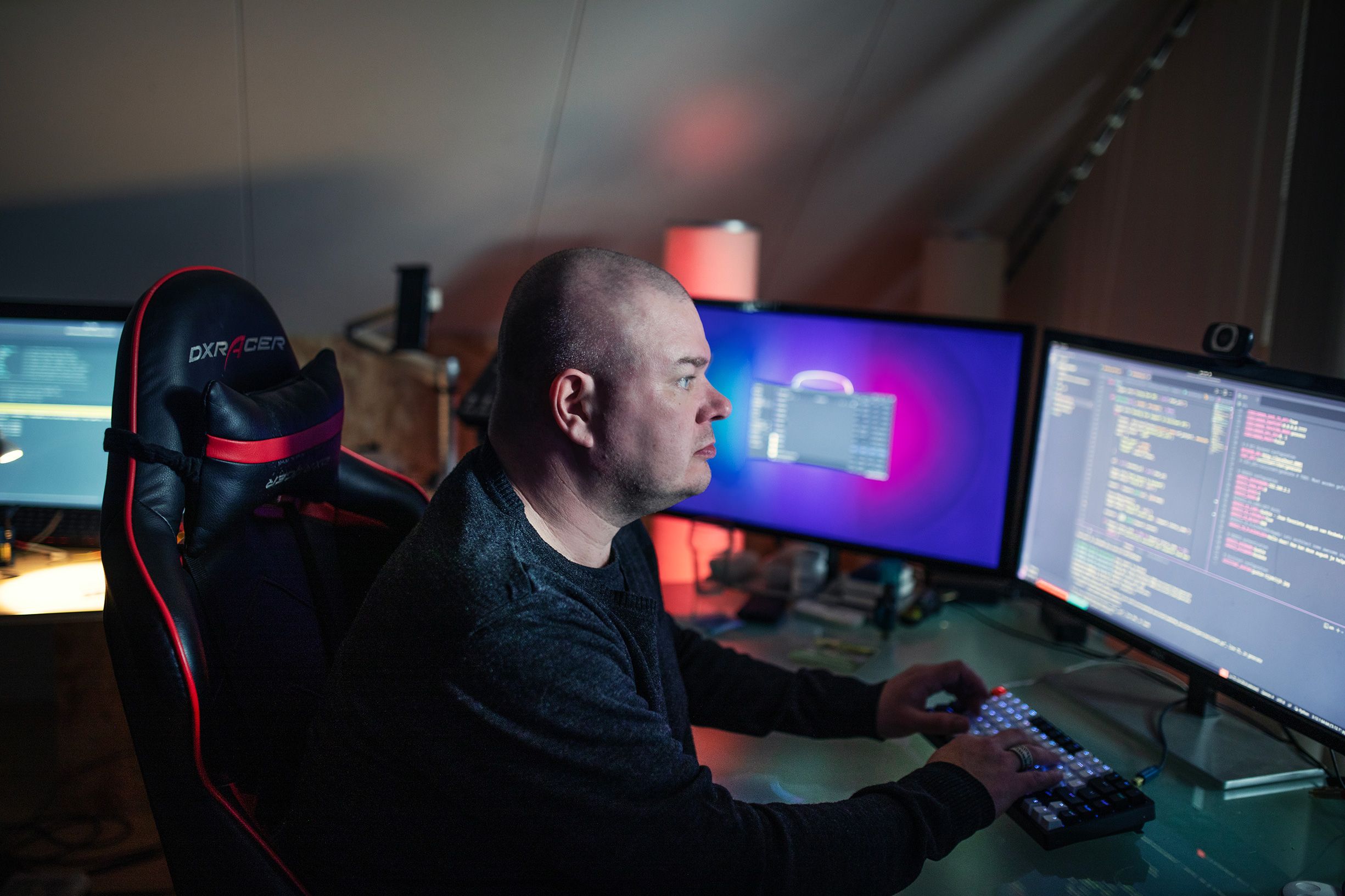 English: Person in dark shirt coding on multiple monitors with programming text visible, sitting in gaming chair with red accents in dimly lit room.