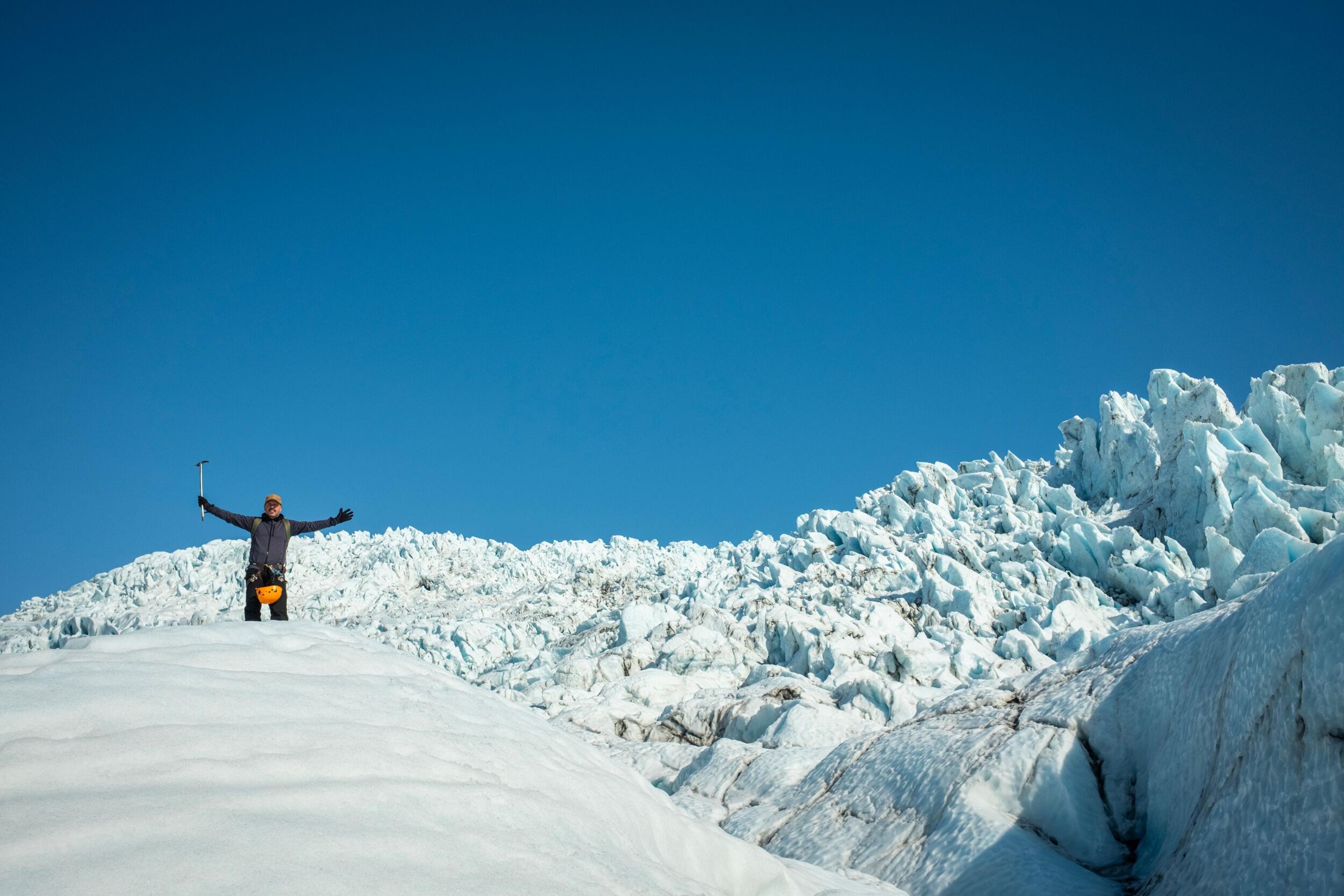 A mountain guide stands with arms outstretched on a snow-covered glacier, with a vast field of jagged blue-white ice formations and a clear blue sky behind them.