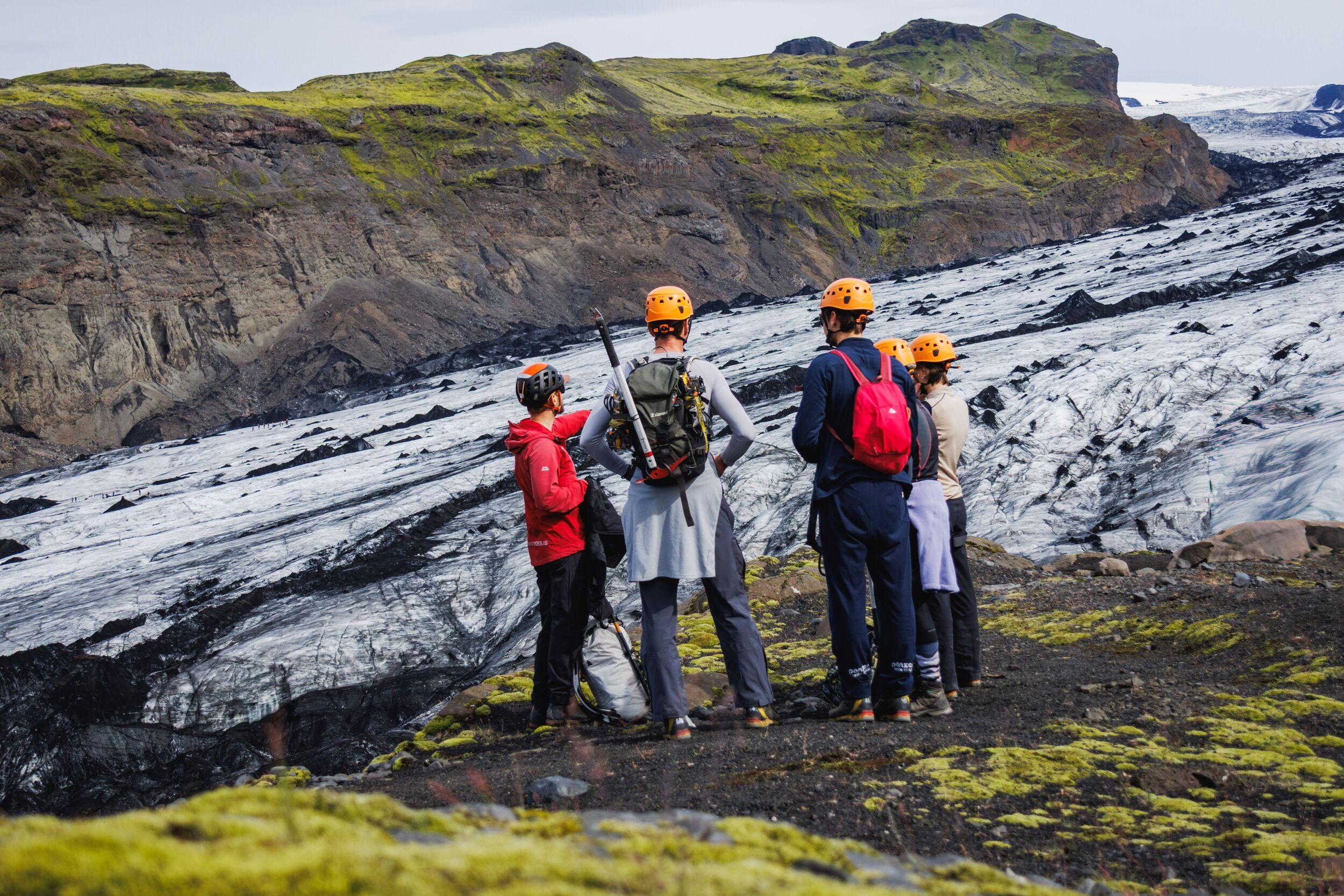 Here's alt text for this one: "A small group of hikers in orange helmets stands on moss-covered ground, looking out over a vast glacier streaked with dark volcanic ash.