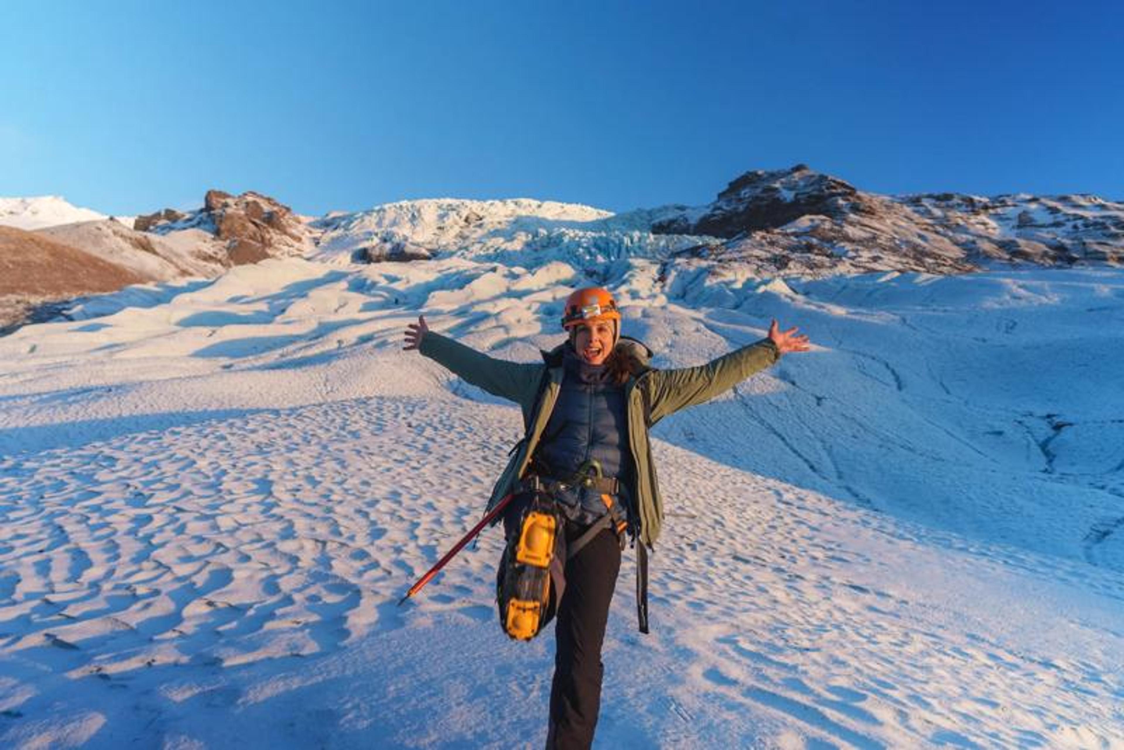 A joyful hiker with arms spread wide and one crampon-fitted boot raised poses on a snowy glacier, with rocky peaks glowing in golden light behind her