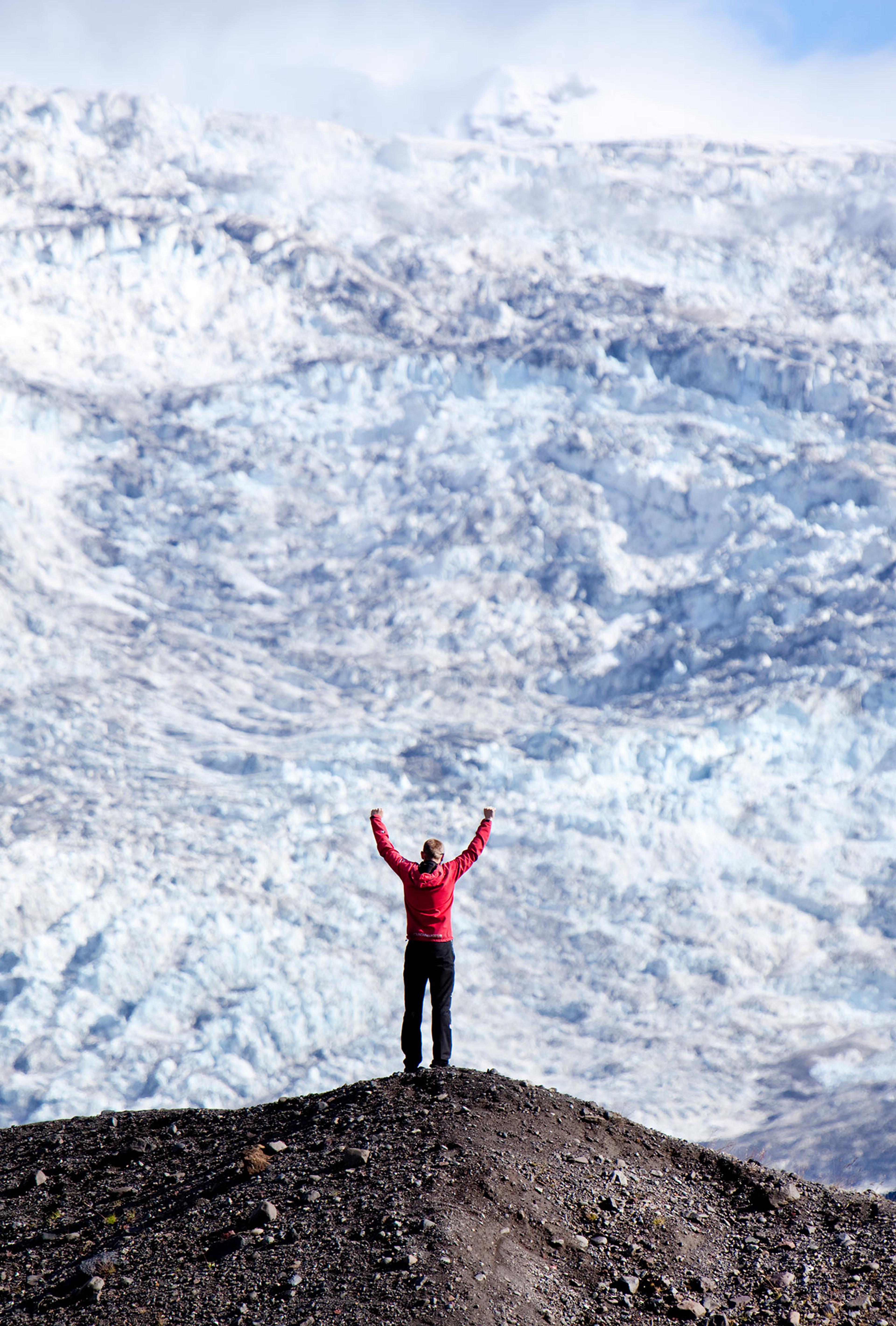 A person in a red jacket on a mountain with their hands in the air. A massive glacier is in front of them.