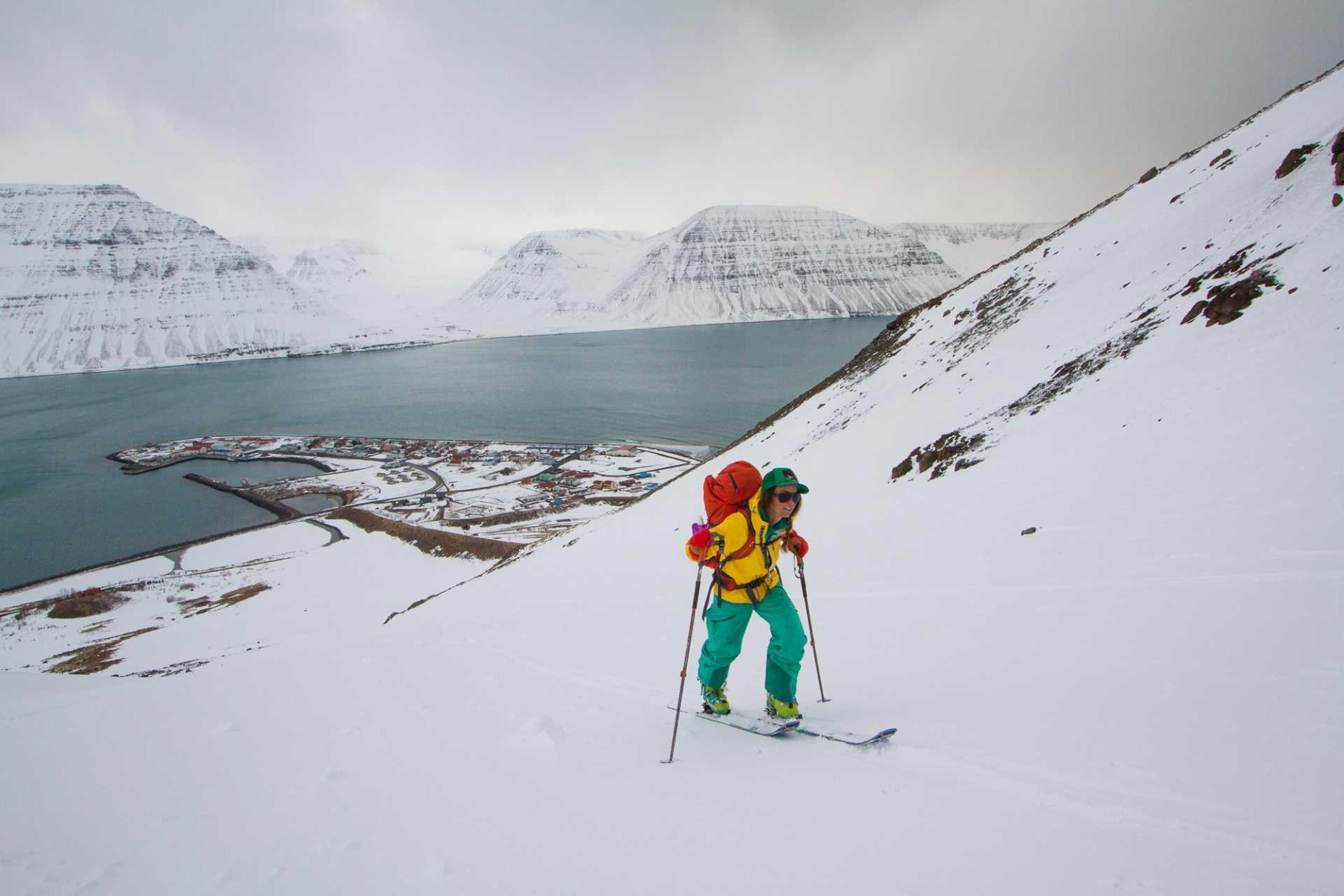 A skier climbs a snowy slope with a backpack, with a picturesque fjord and a small village in the background.