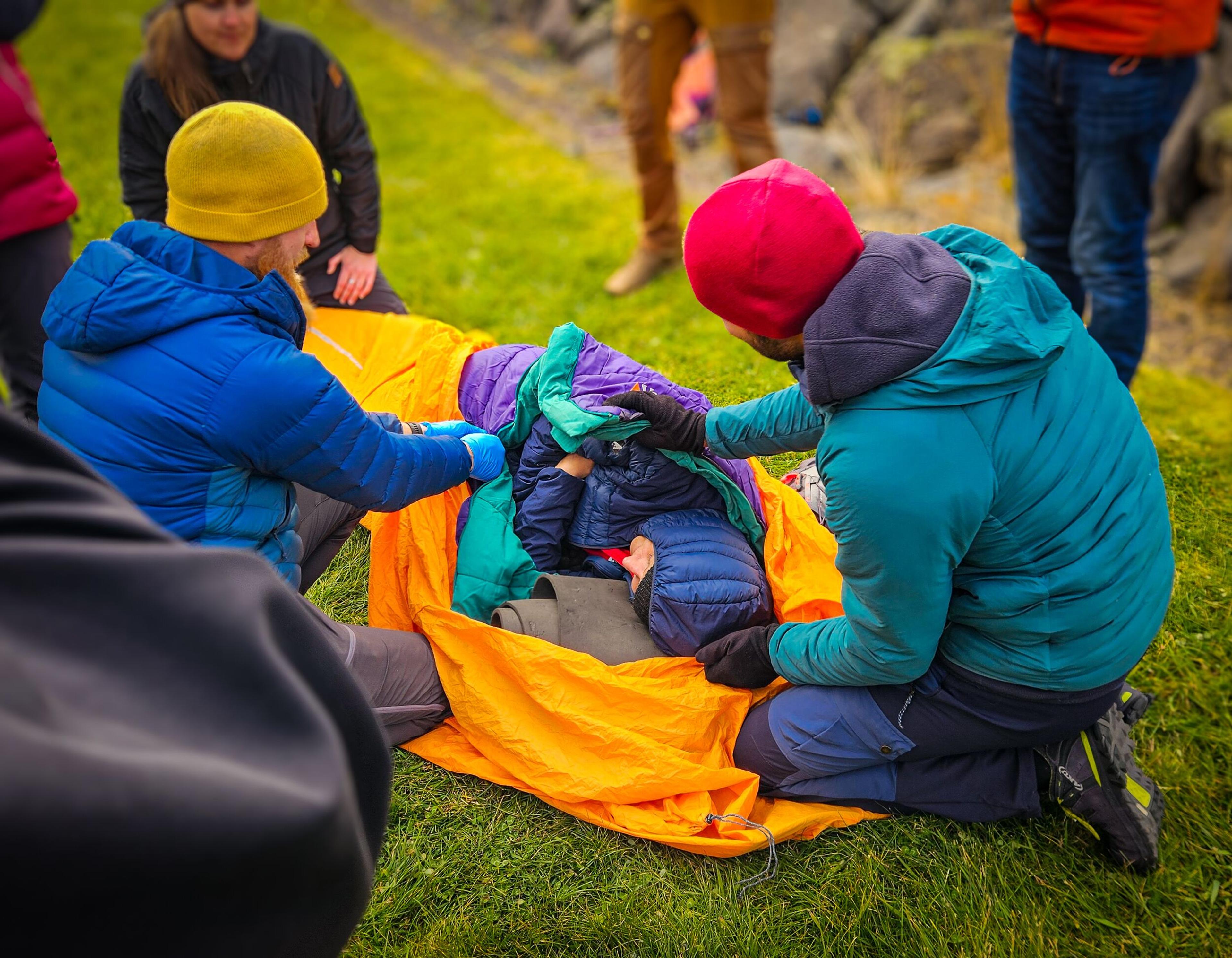 a group of people in a wilderness setting practicing a first responder or emergency care technique. A person is wrapped in colorful insulated blankets