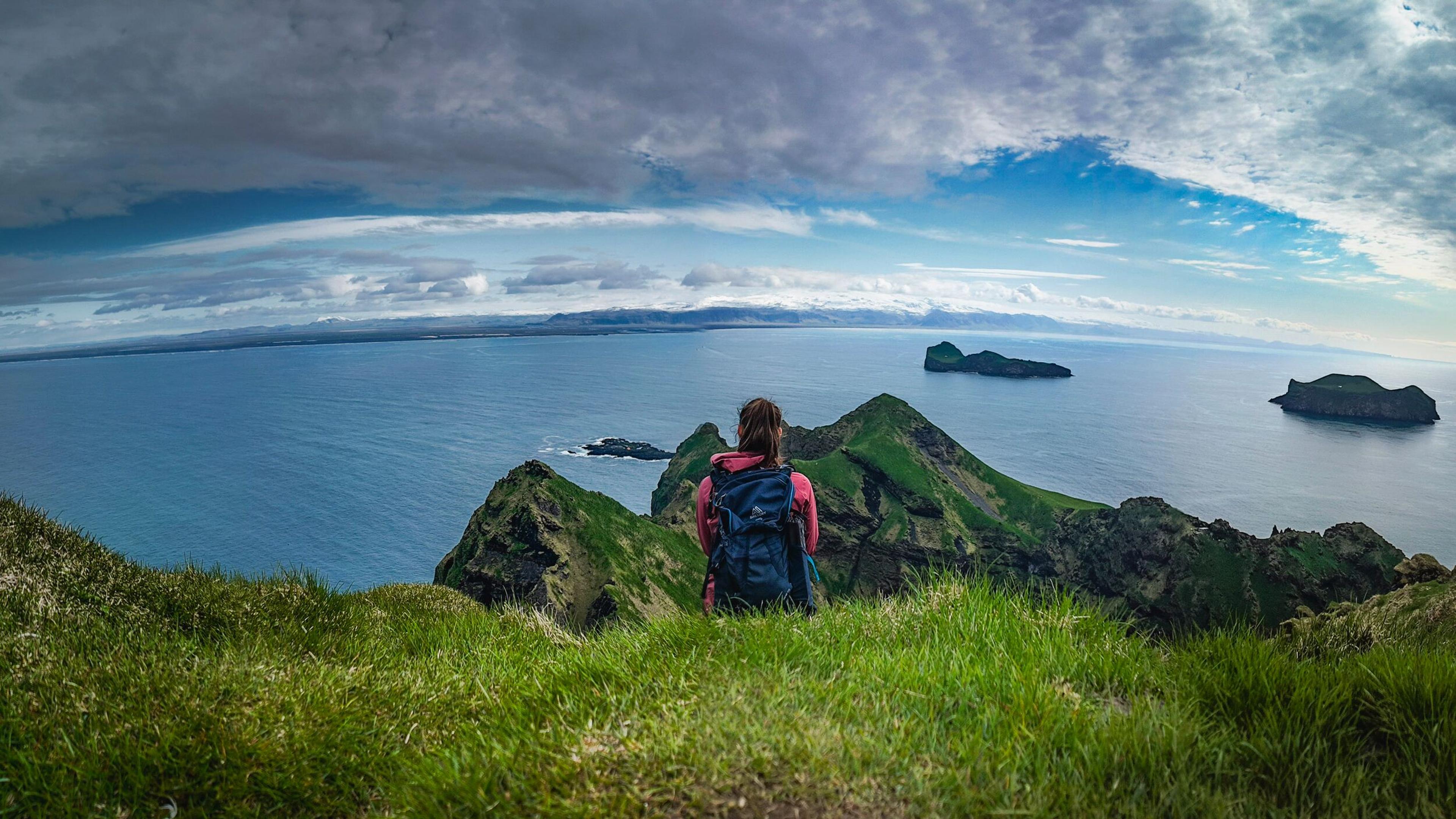 A woman hiker sat at the top of a grassy mountain in Iceland overlooking the North Atlantic Ocean