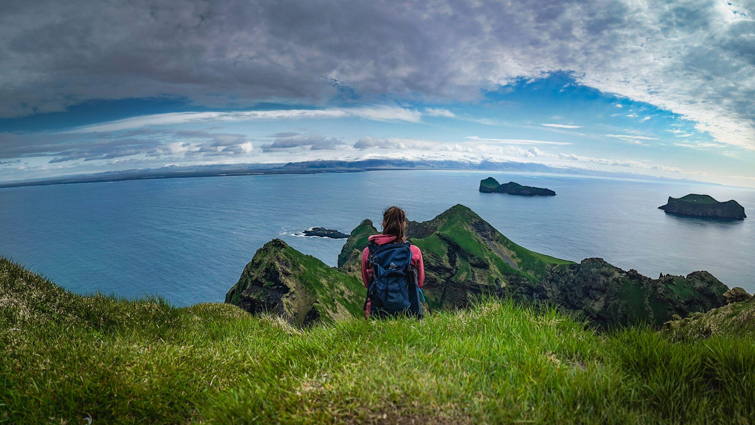 A woman hiker sat at the top of a grassy mountain in Iceland overlooking the North Atlantic Ocean