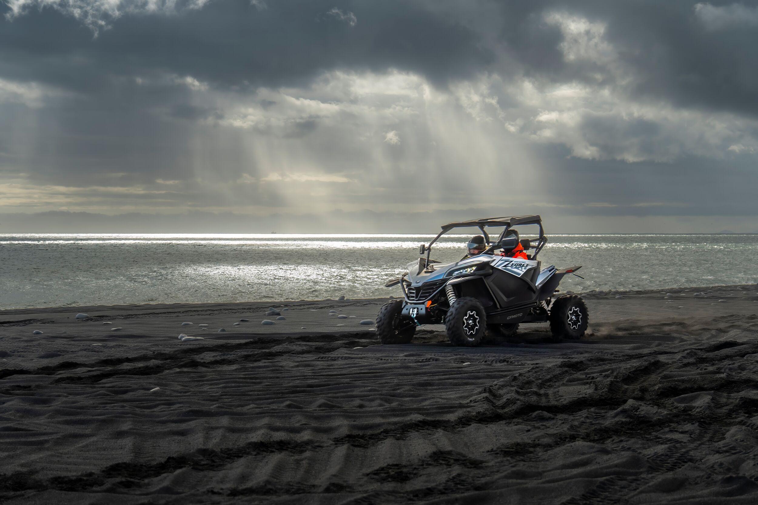A buggy speeds along a black sand beach, with dramatic sun rays breaking through the clouds and reflecting on the ocean.