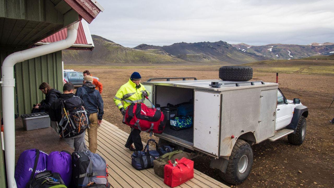 A group of hikers unloads gear and luggage from a white off-road vehicle parked at a remote mountain hut along the Laugavegur trail in Iceland.