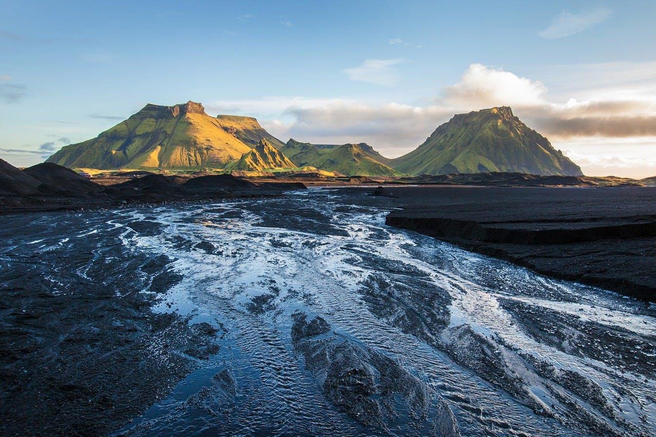 Green mountains in the distance  rising over black sands with a small river running over the sand