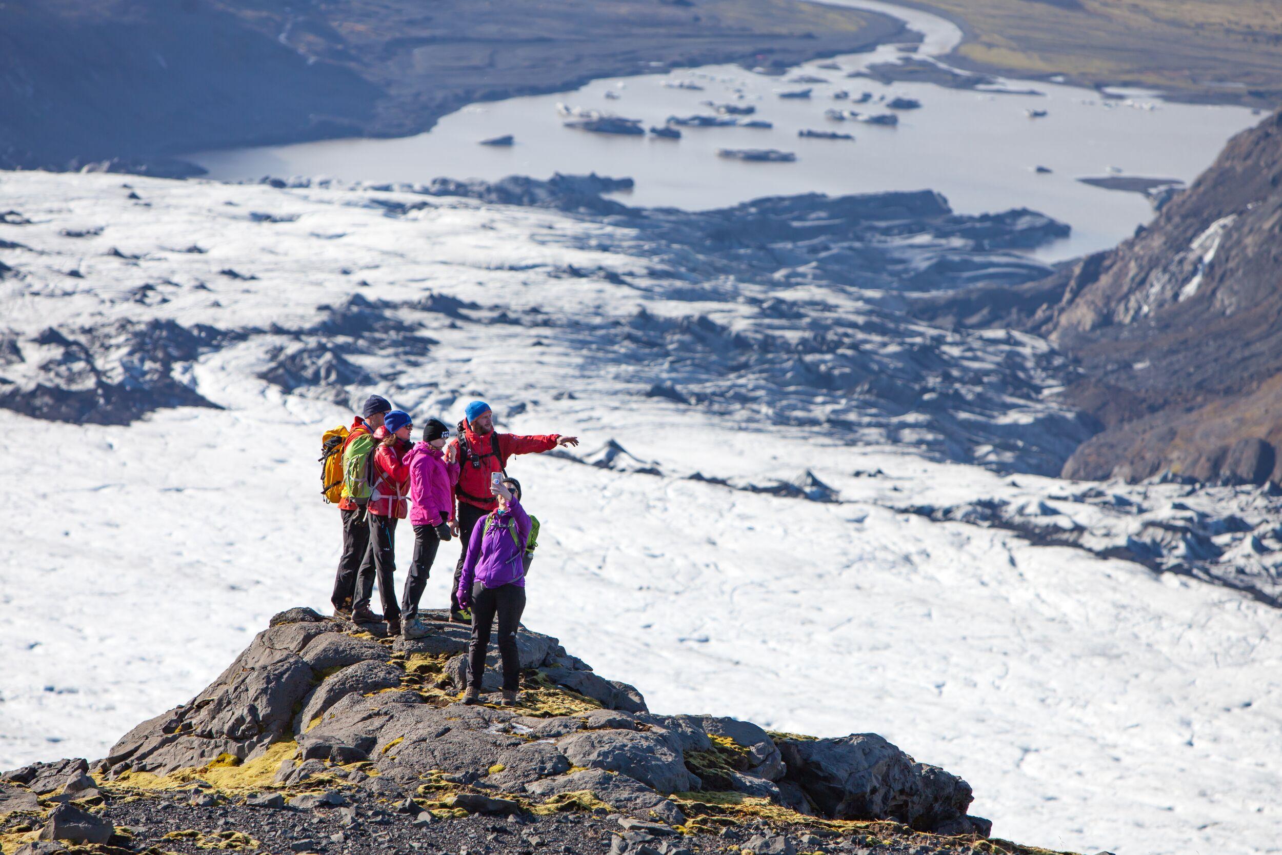 5 hikers standing on a cliff above a glacier and lagoon below