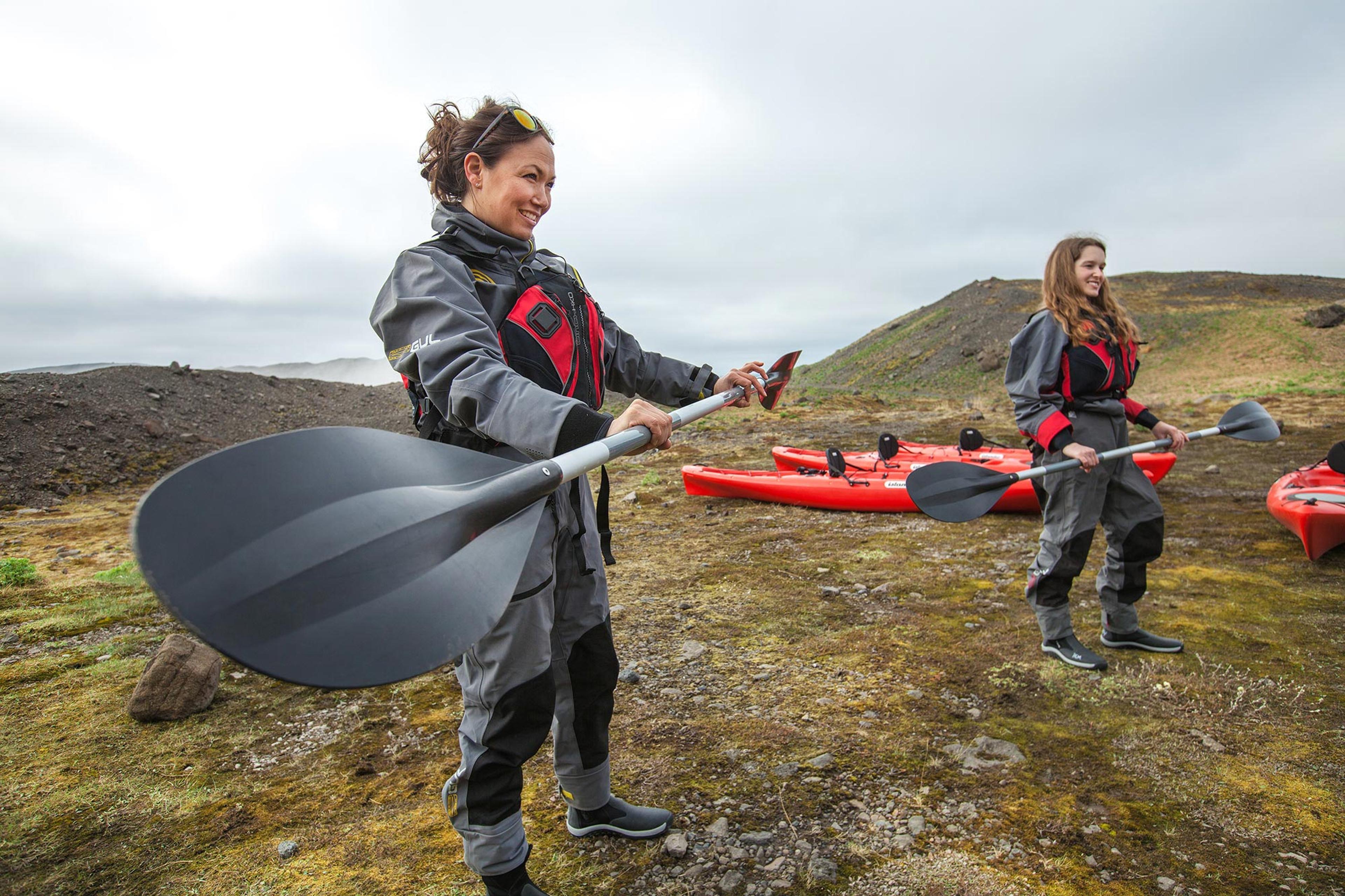 Two women in drysuits holding kayaking paddles out in Icelandic nature.