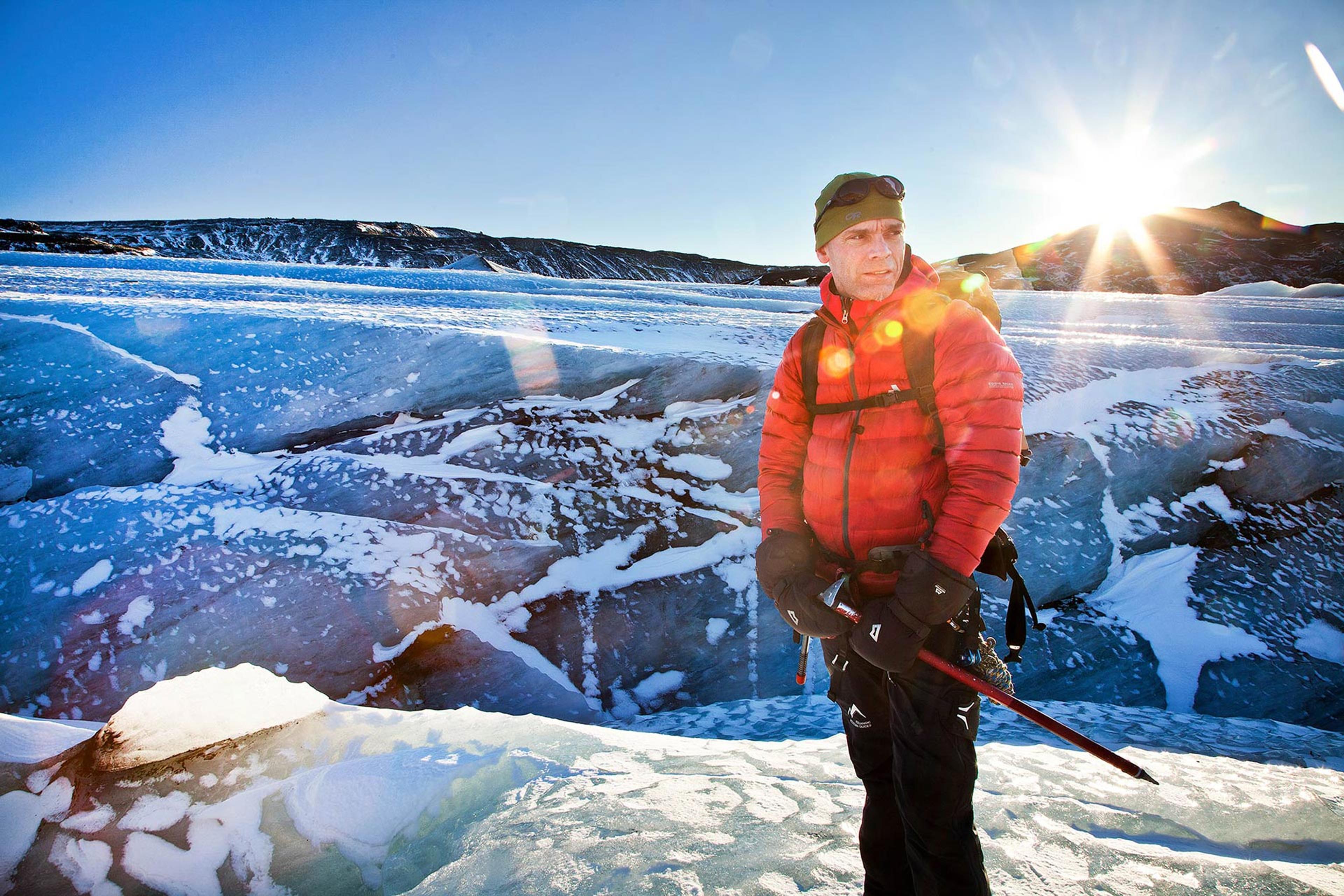 A man in red holding an ice axe standing on Sólheimajökull while the sun sets behind him
