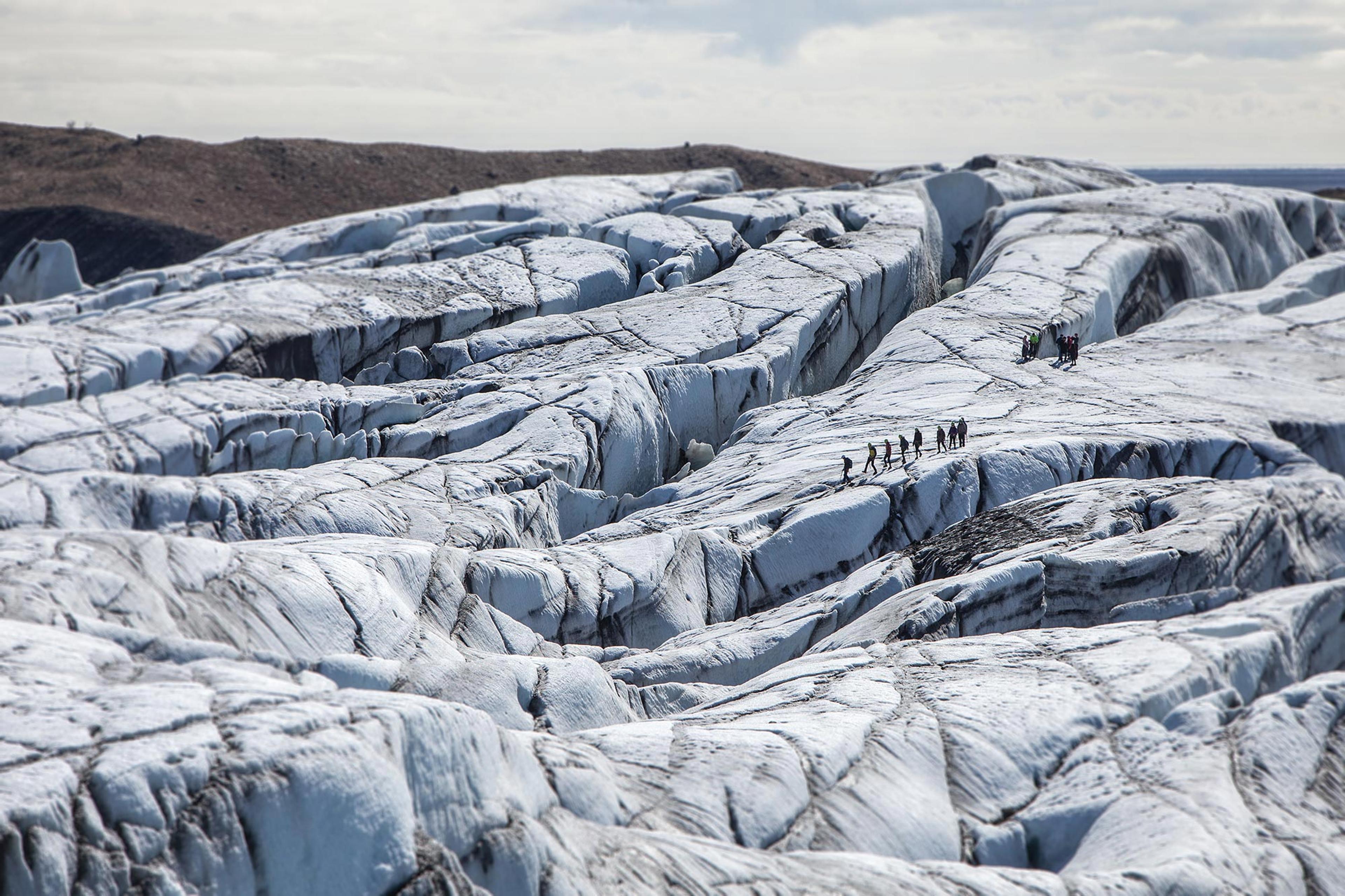 A vast glacial landscape of blue, white and black. There is a group glacier hiking in the distance.
