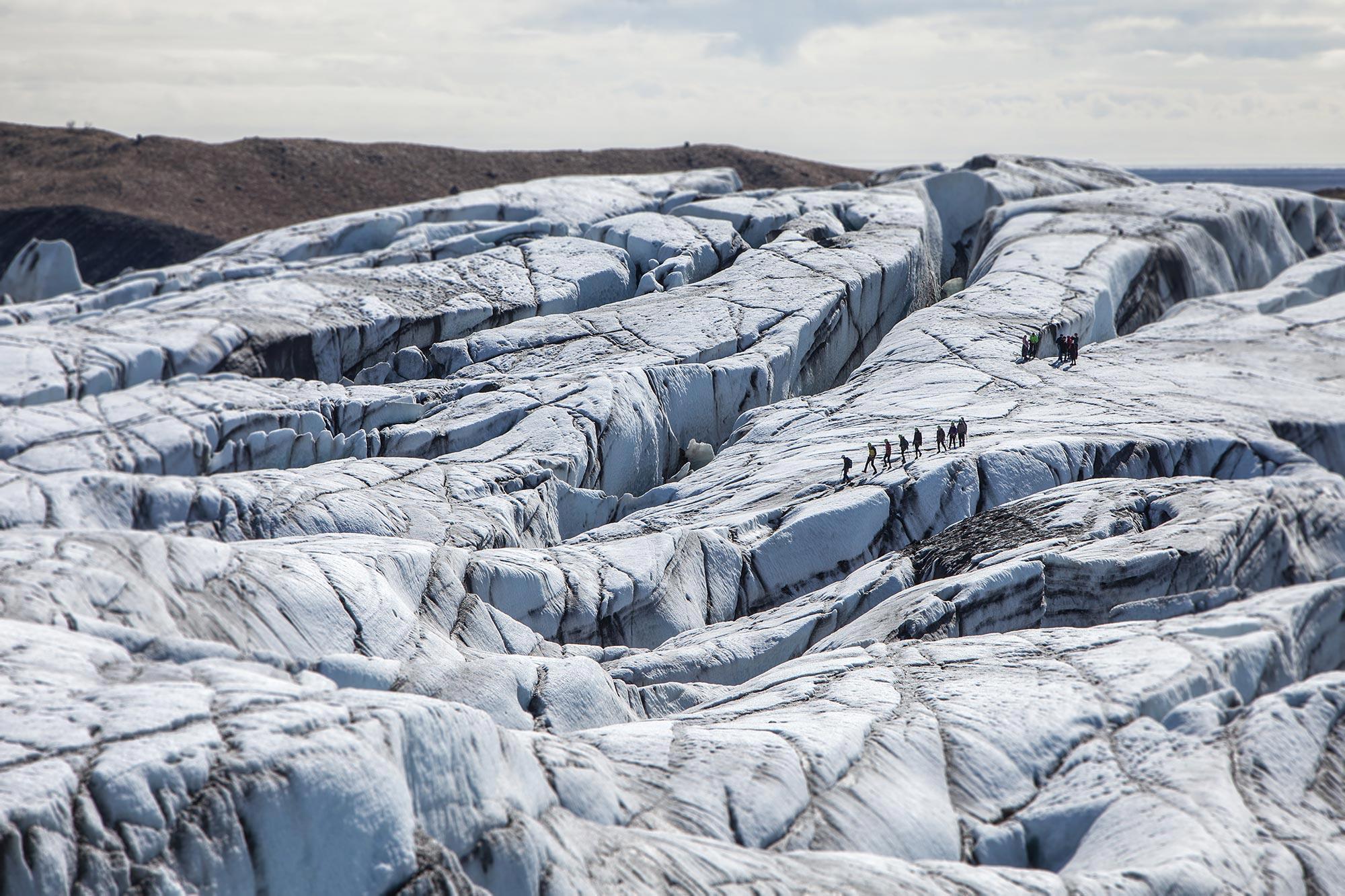 A vast glacial landscape of blue, white and black. There is a group glacier hiking in the distance.