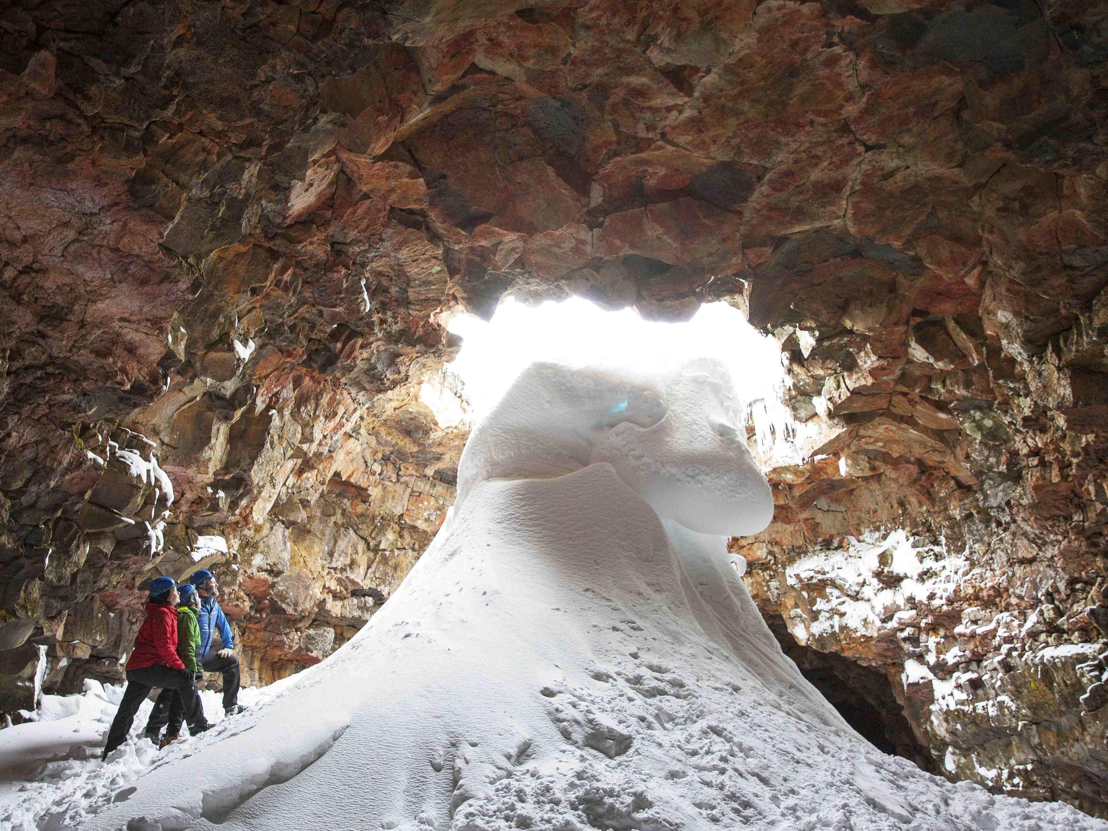 A large snow formation by the entrance of the cave and three people looking up to the entrance.