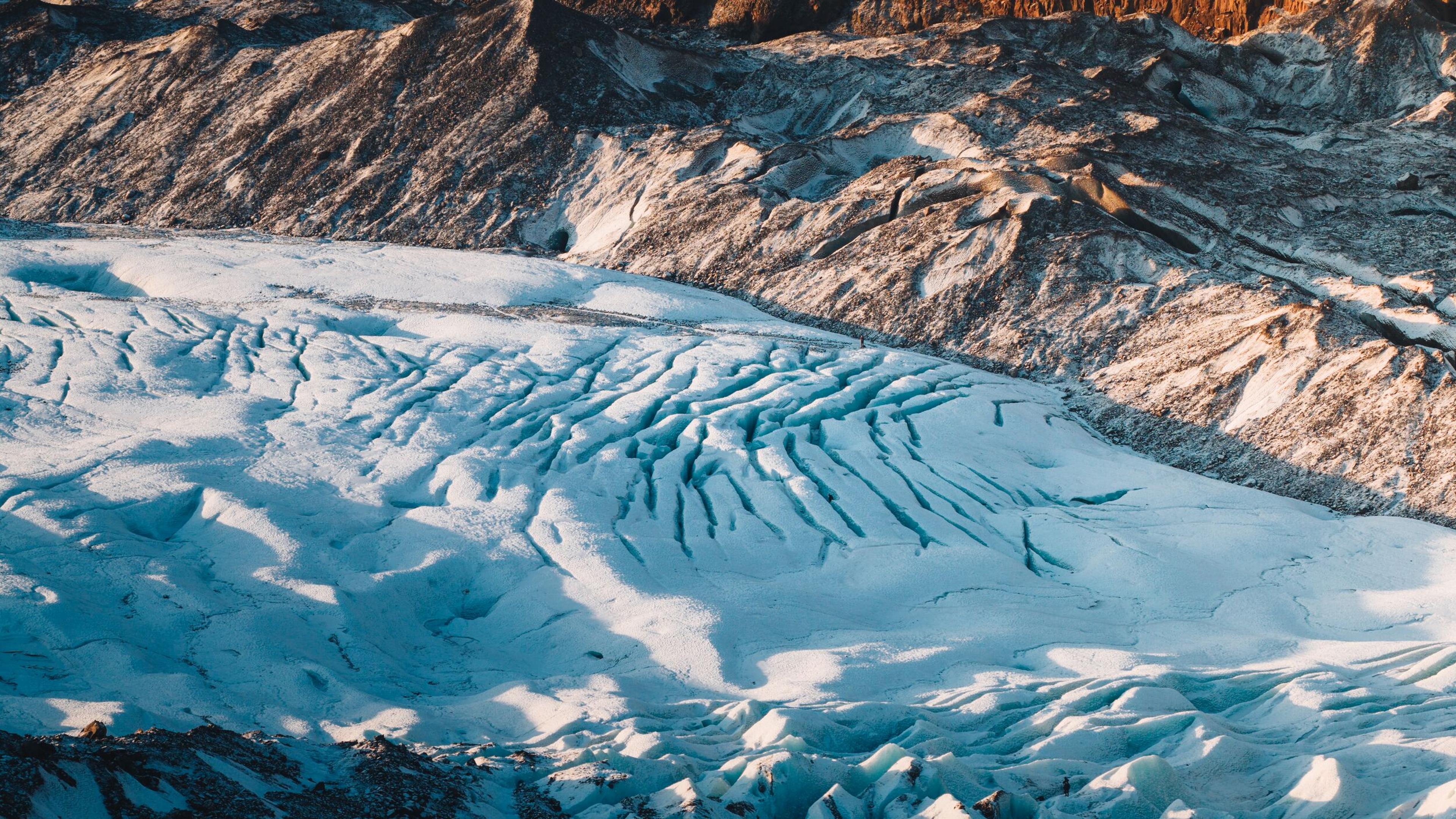 "Aerial view of a glacier tongue with a fan of deep blue crevasses cutting through the ice, bordered by dark volcanic rock formations