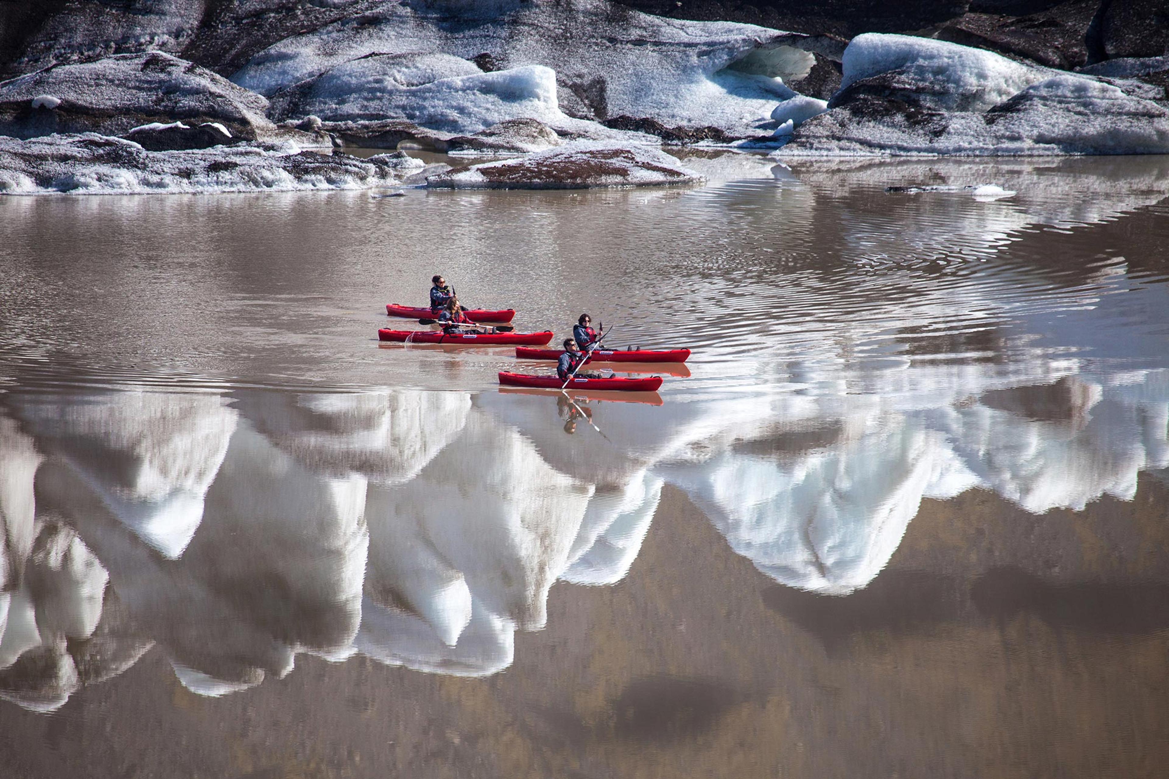 Four people in red kayaks gliding on Solheimajokull’s glacier lagoon with icebergs reflecting in the water.