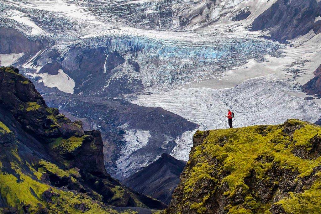 I hiker standing on a green cliff overlooking a thrilling icefall of a glacier
