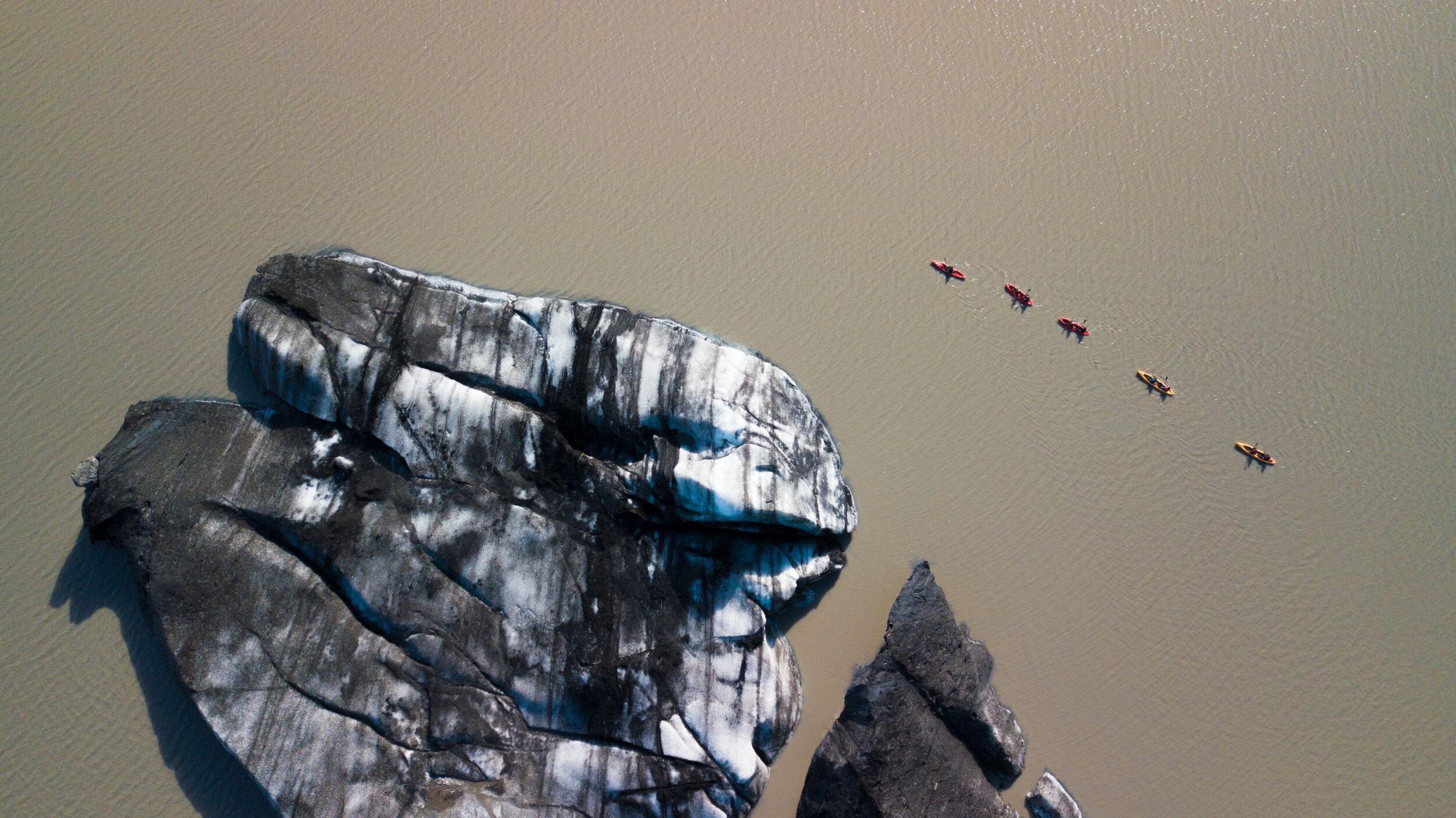 Aerial view of a large, dark-and-white glacier ice formation in a glacial lake, with five small kayaks paddling nearby.