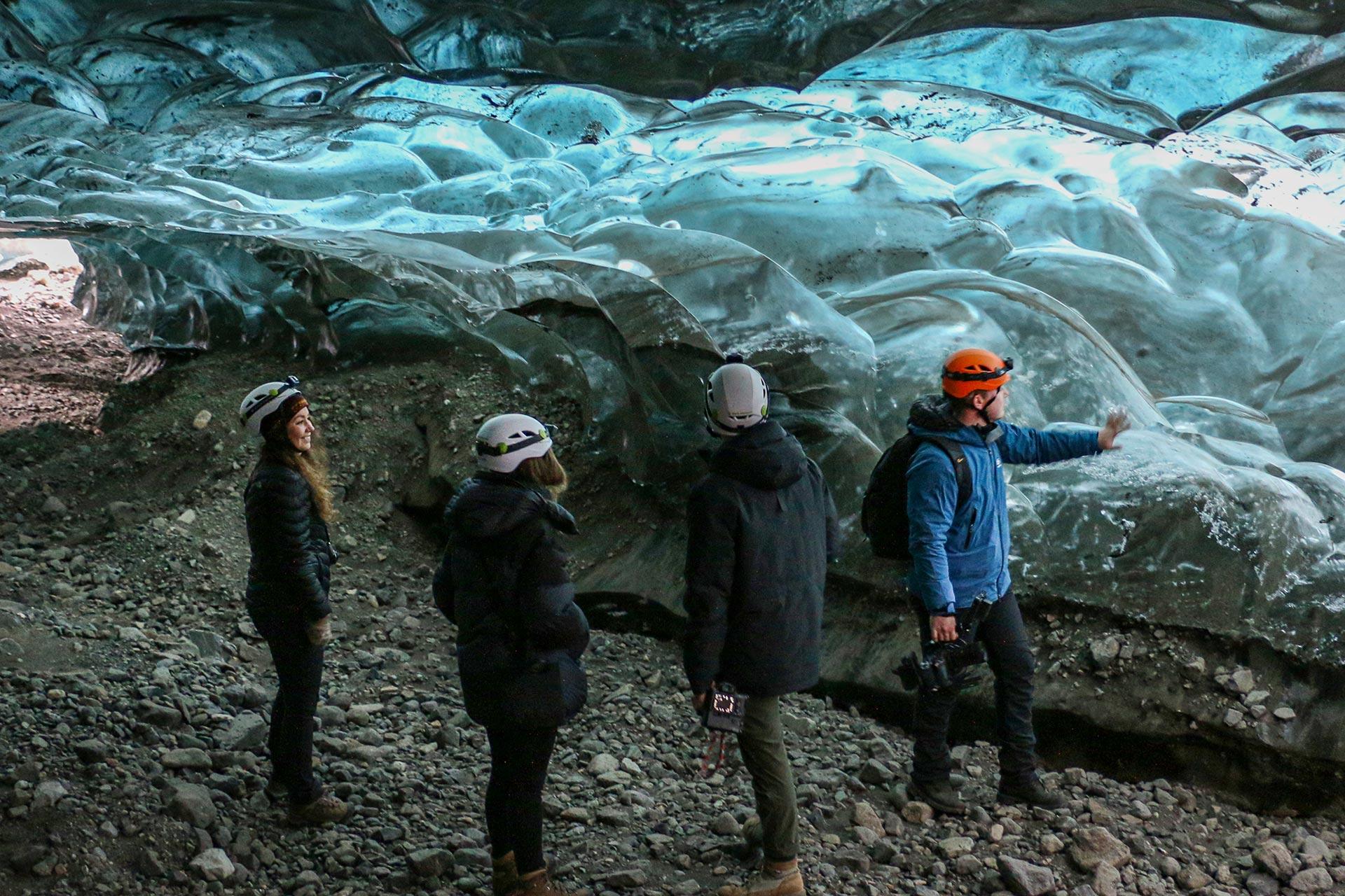 Three individuals are standing in a dimly lit ice cave, observing the wavy patterns of the blue and white ice wall in front of them. One person, wearing a bright orange helmet, points towards the ice structure, possibly providing explanations or instructions to the others.