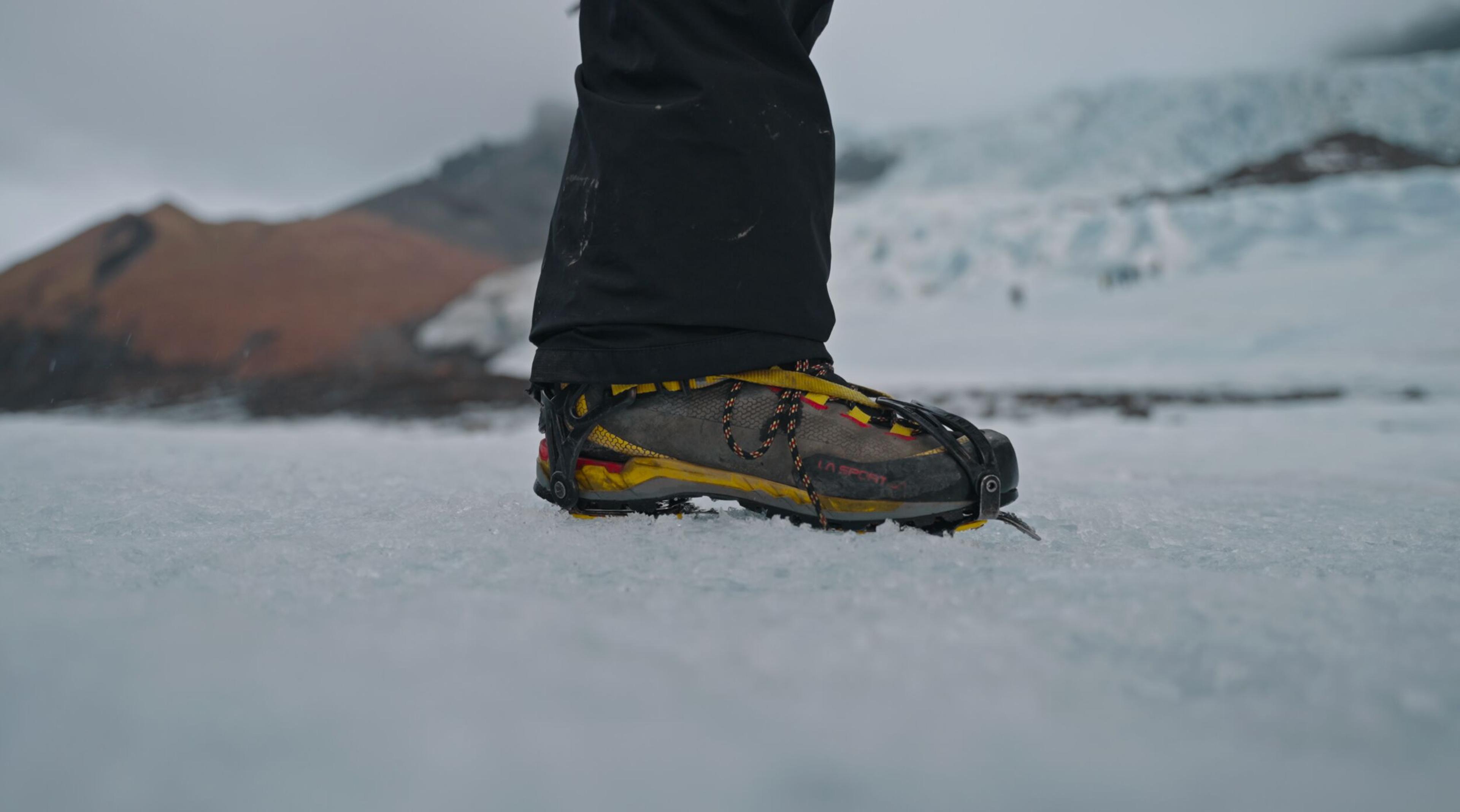 Close-up of a hiking boot fitted with a crampon attachment, standing on icy glacier terrain with a blue ice formation blurred in the background