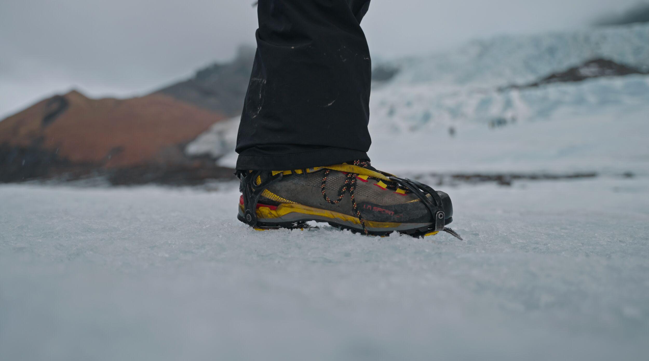 Close-up of a hiking boot fitted with a crampon attachment, standing on icy glacier terrain with a blue ice formation blurred in the background