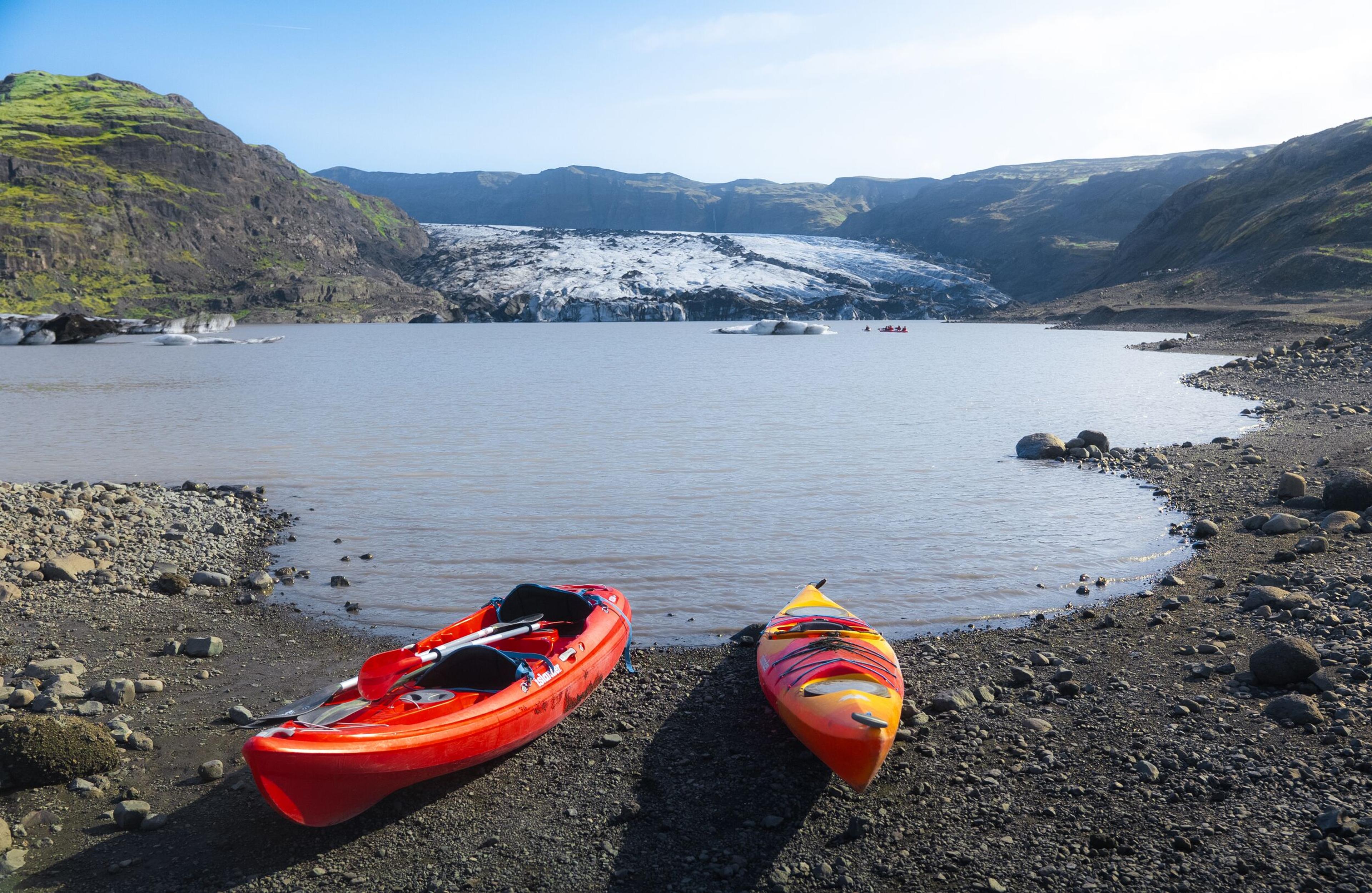 Two colorful kayaks rest on a rocky shoreline beside a calm glacial lake, with floating ice and a large glacier surrounded by rugged, green-topped mountains in the background.