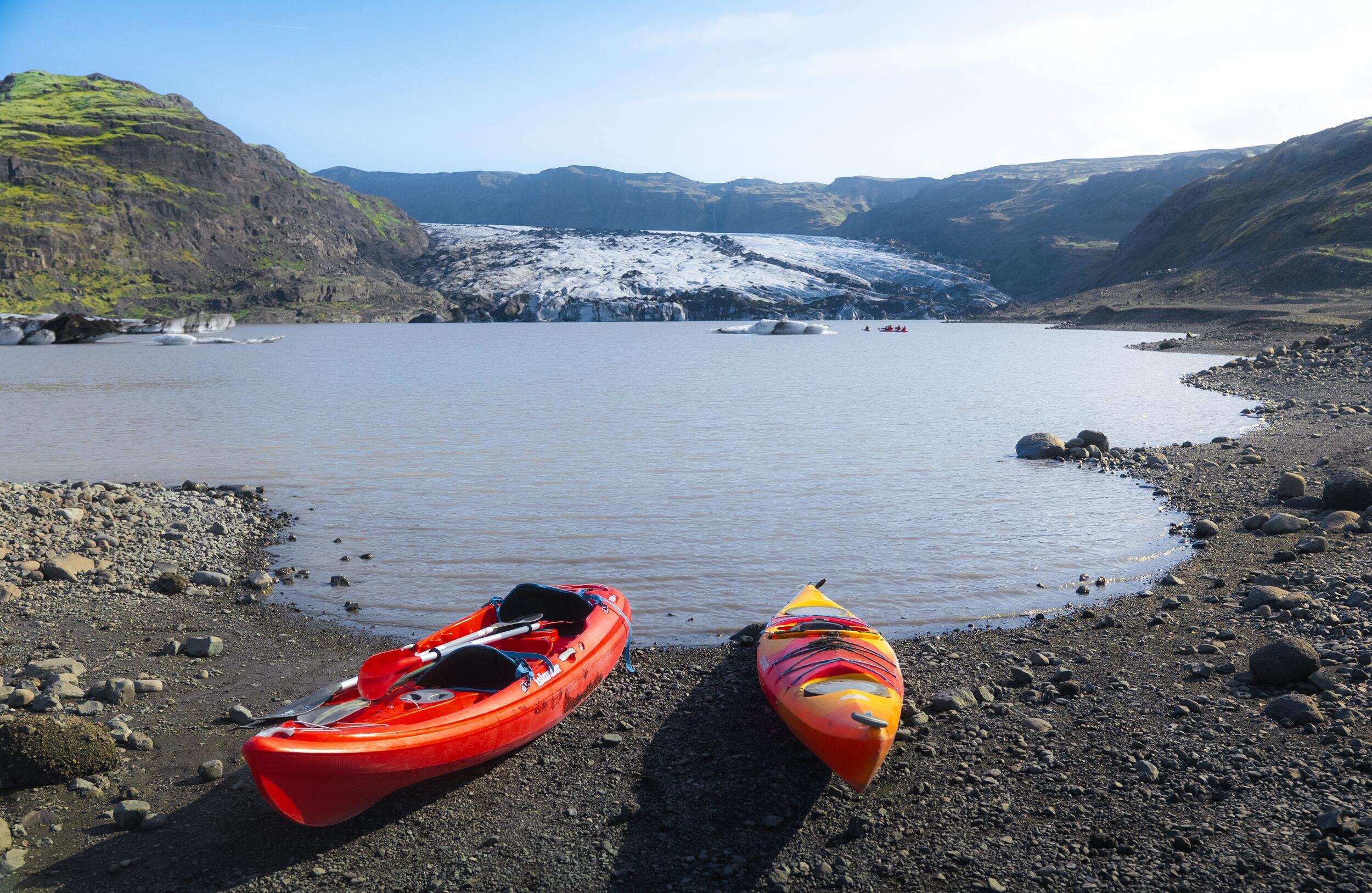 Two colorful kayaks rest on a rocky shoreline beside a calm glacial lake, with floating ice and a large glacier surrounded by rugged, green-topped mountains in the background.