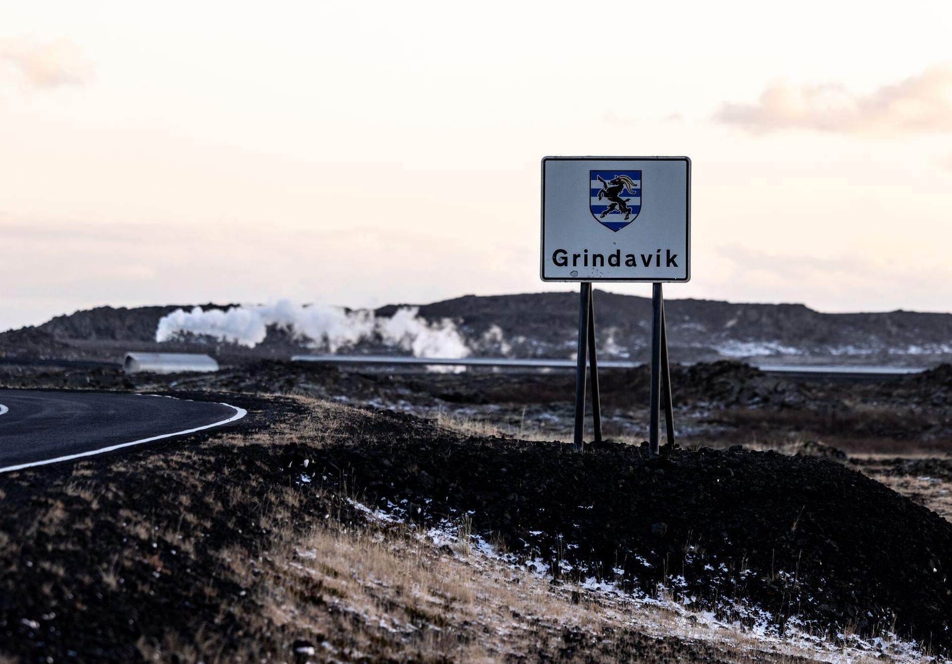 A road sign with the Grindavik town name and emblem stands amidst a rugged volcanic landscape, with steam rising from geothermal vents in the background. The winding road and barren terrain emphasize the remote and dramatic setting of this Icelandic region.