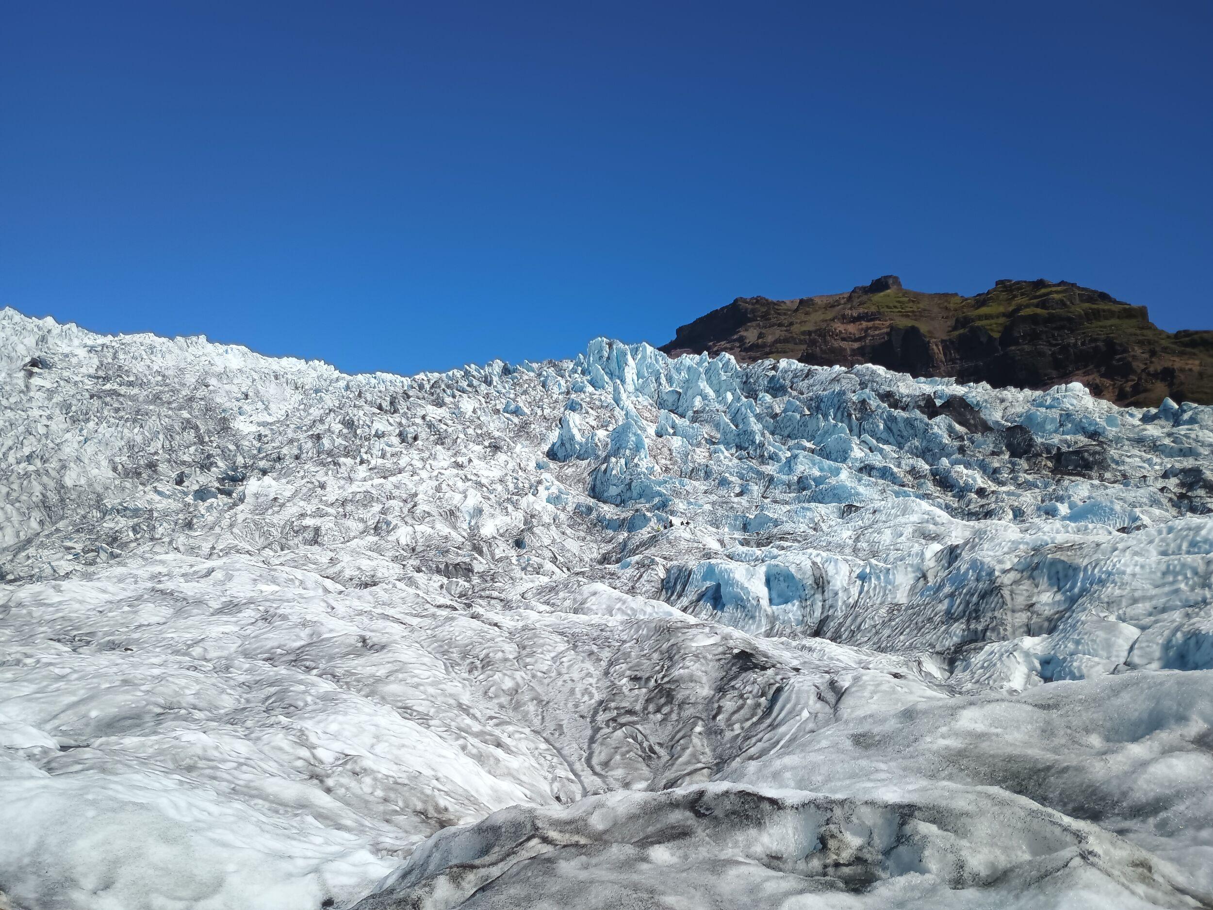 A glacier tongue crawling down a hill with cragged blue edges and ash formations